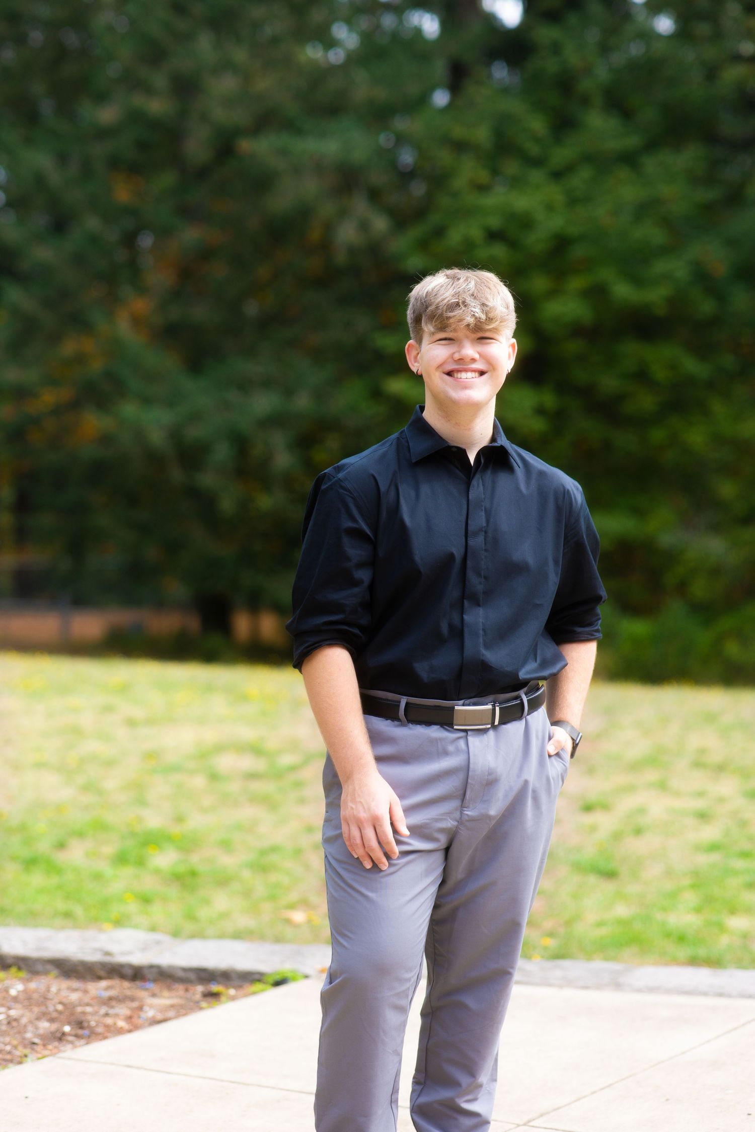 Someone wearing a black collared shirt and gray pants stands outdoors on a walkway surrounded by greenery.