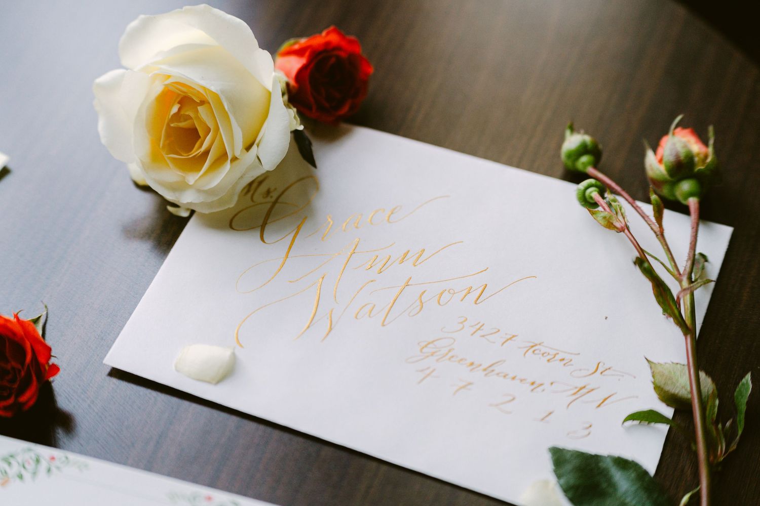Flat lay of a wedding invitation suite framed by lush florals in red, blush, and cream tones, with delicate hand lettering and detailing