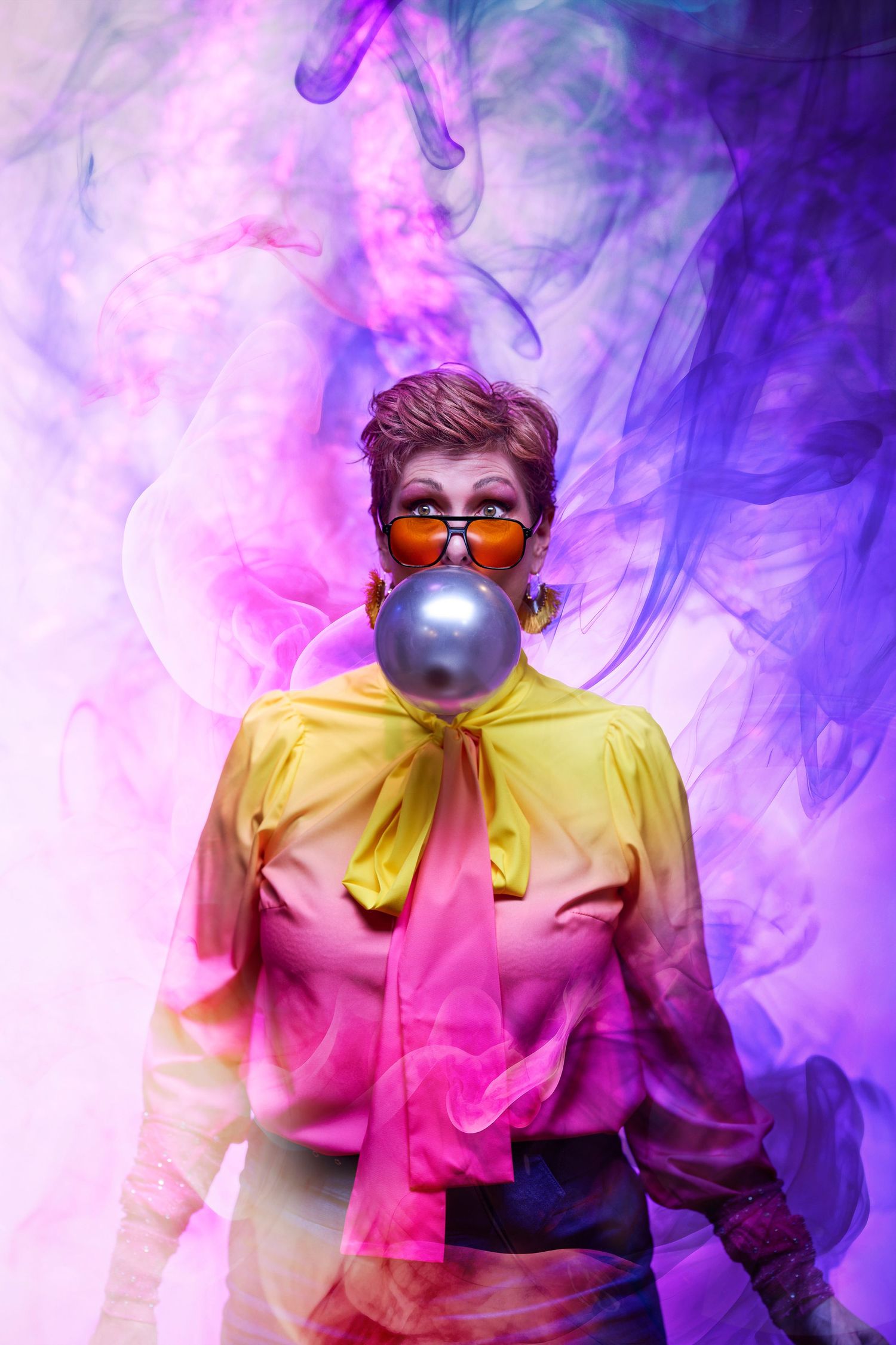 Person in yellow and pink gradient shirt blowing bubblegum against colorful smoke background with sunglasses.