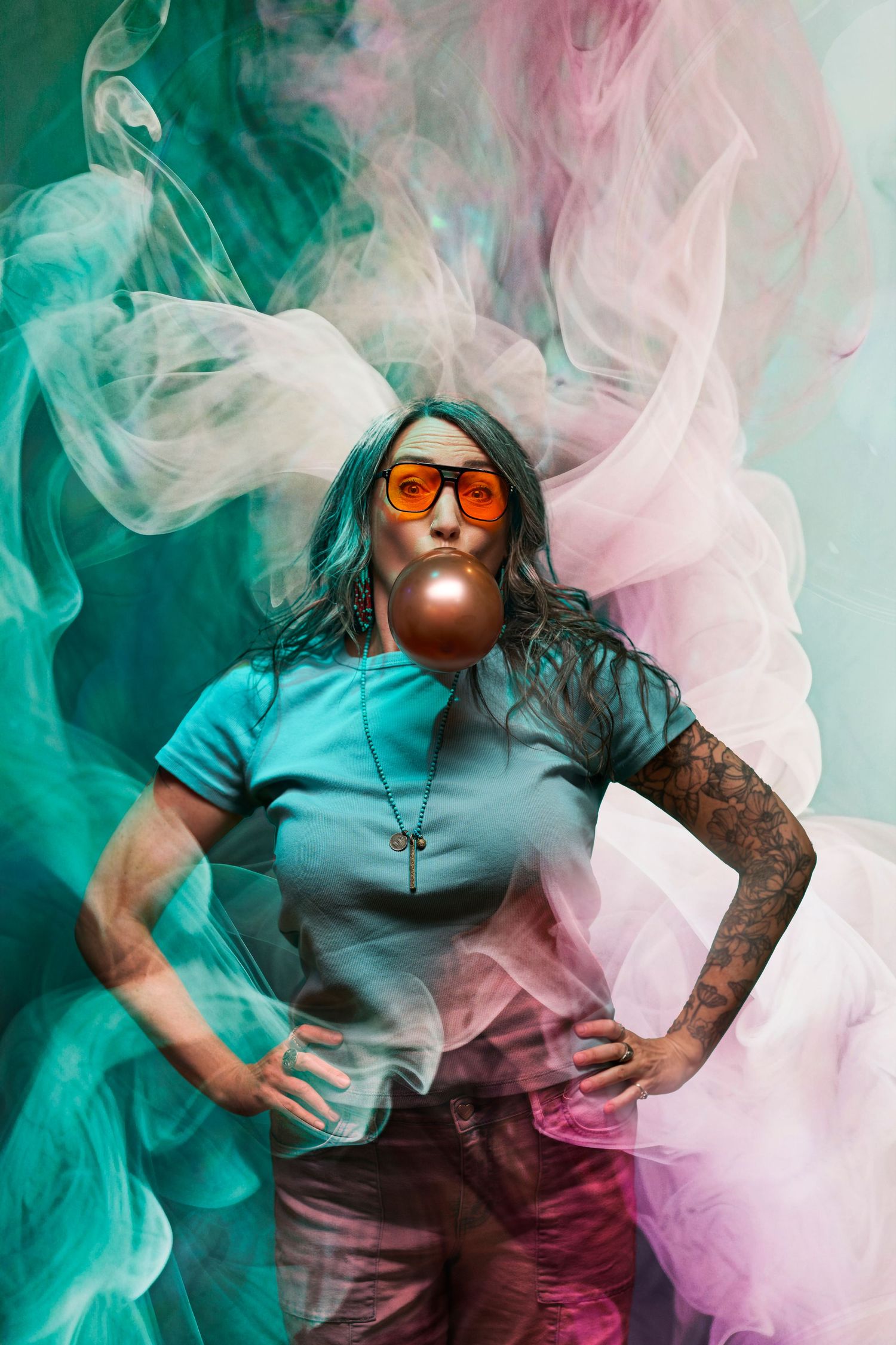 Stylish portrait with orange sunglasses and tattoos surrounded by swirling teal and pink smoke against dark background.