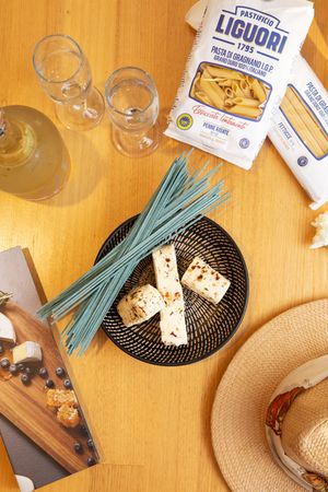 Overhead view of pasta ingredients with blue noodles, cheese, wine glasses, and a straw hat on wooden table.