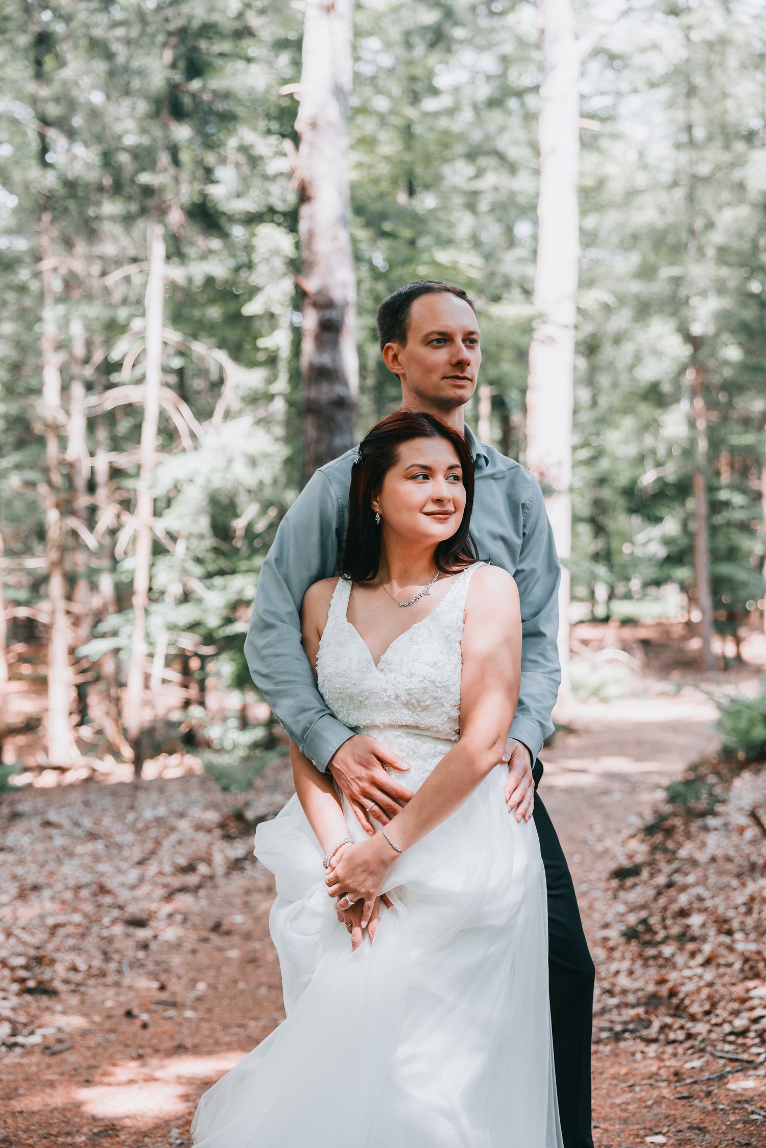A romantic couple in wedding attire embraces while standing in a sunlit forest setting.
