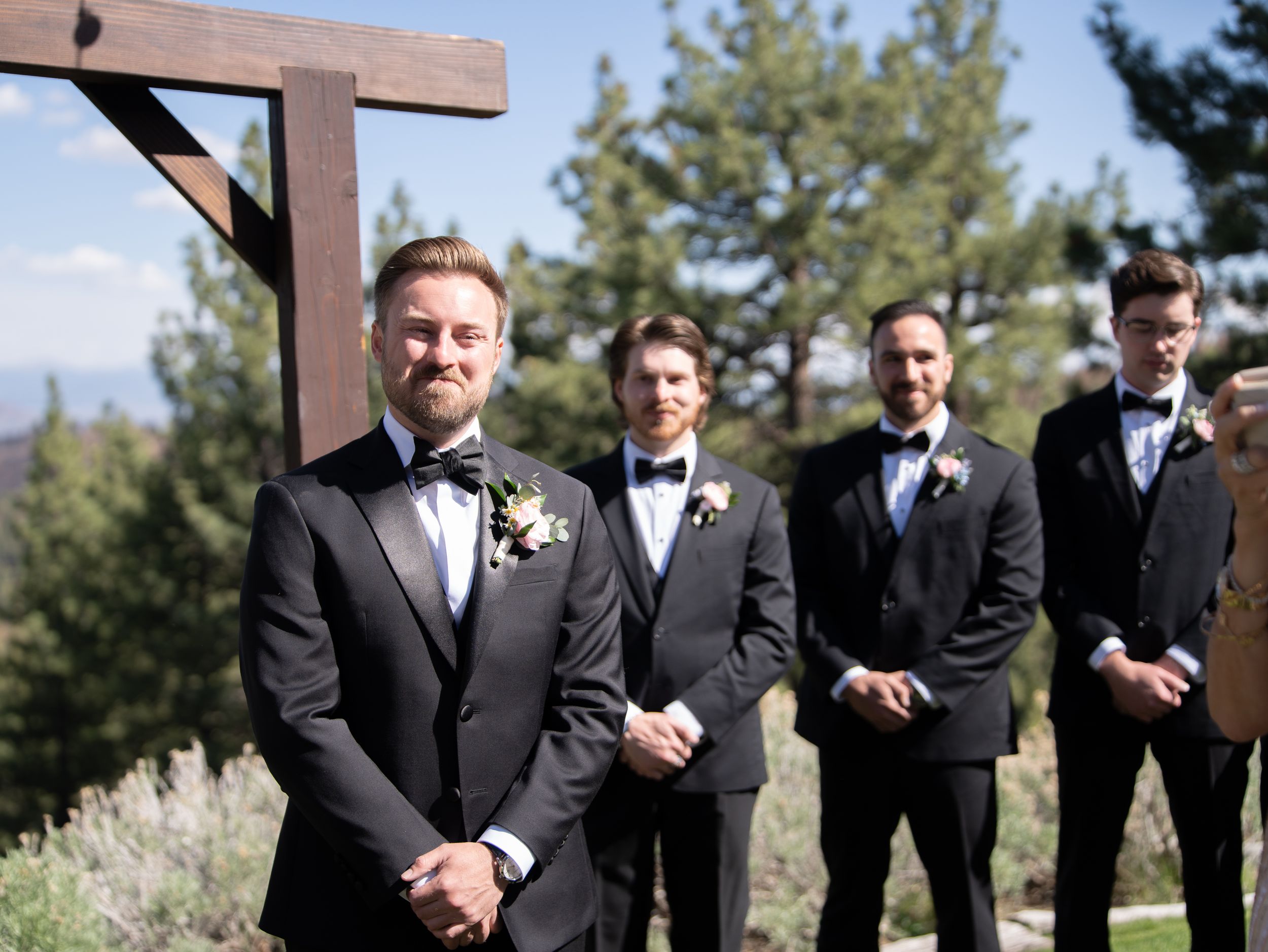 A group of formally dressed groomsmen in black suits stand outdoors for wedding party photos.