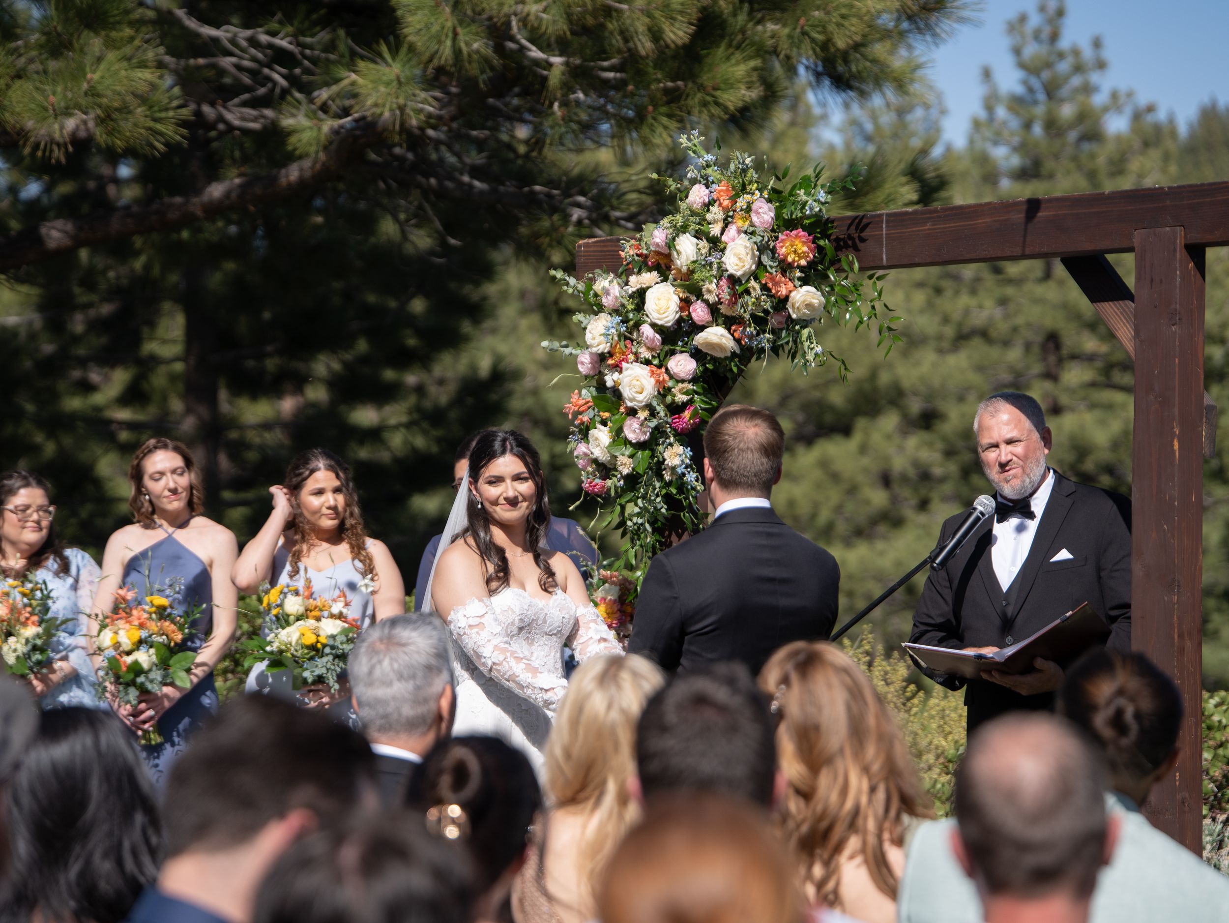 An outdoor wedding ceremony under a wooden arch adorned with pink and white floral arrangements in a pine forest setting.