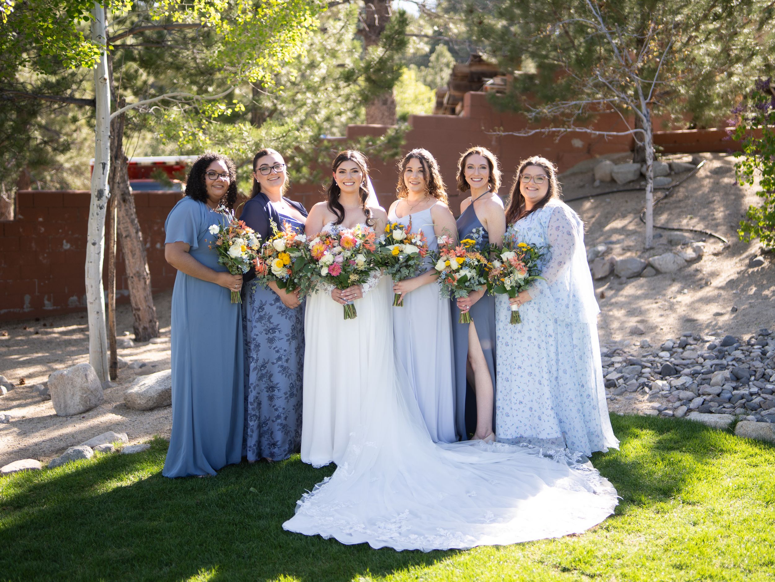Wedding bridal party in light blue and white dresses pose together in a sunny garden setting.
