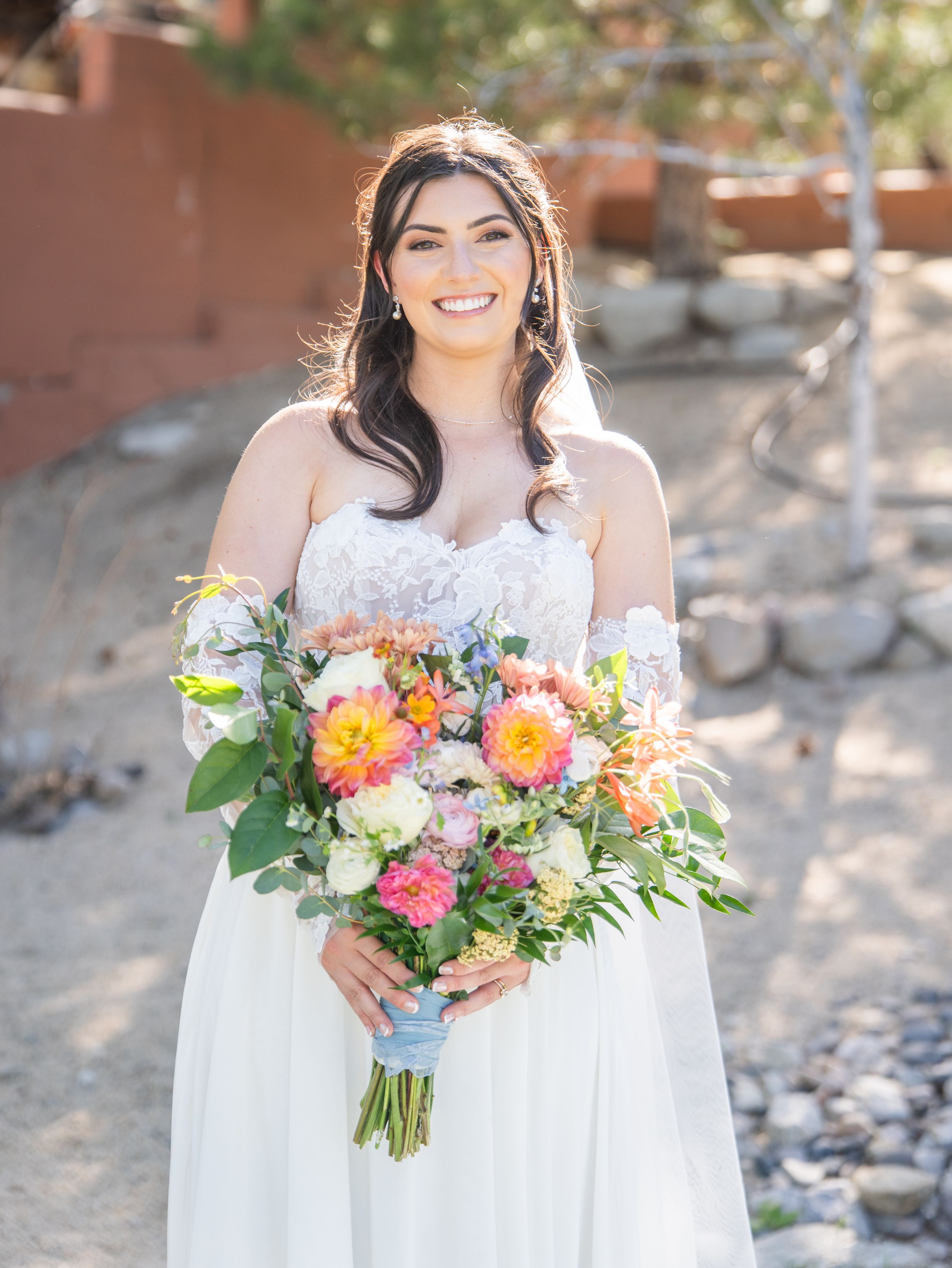 A person in a white dress holds a vibrant wedding bouquet with orange, pink and white flowers against a natural backdrop.