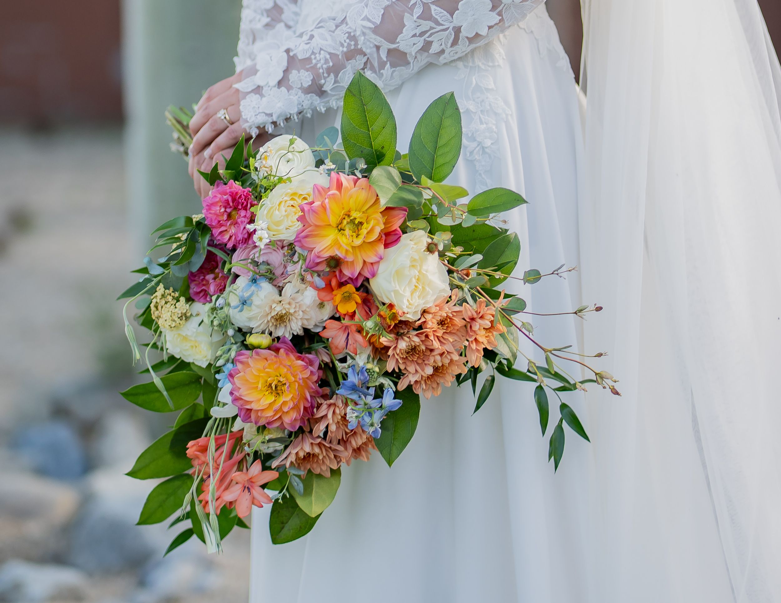 Vibrant bridal bouquets with orange, pink and blue flowers arranged with greenery against flowing white wedding dress fabric.