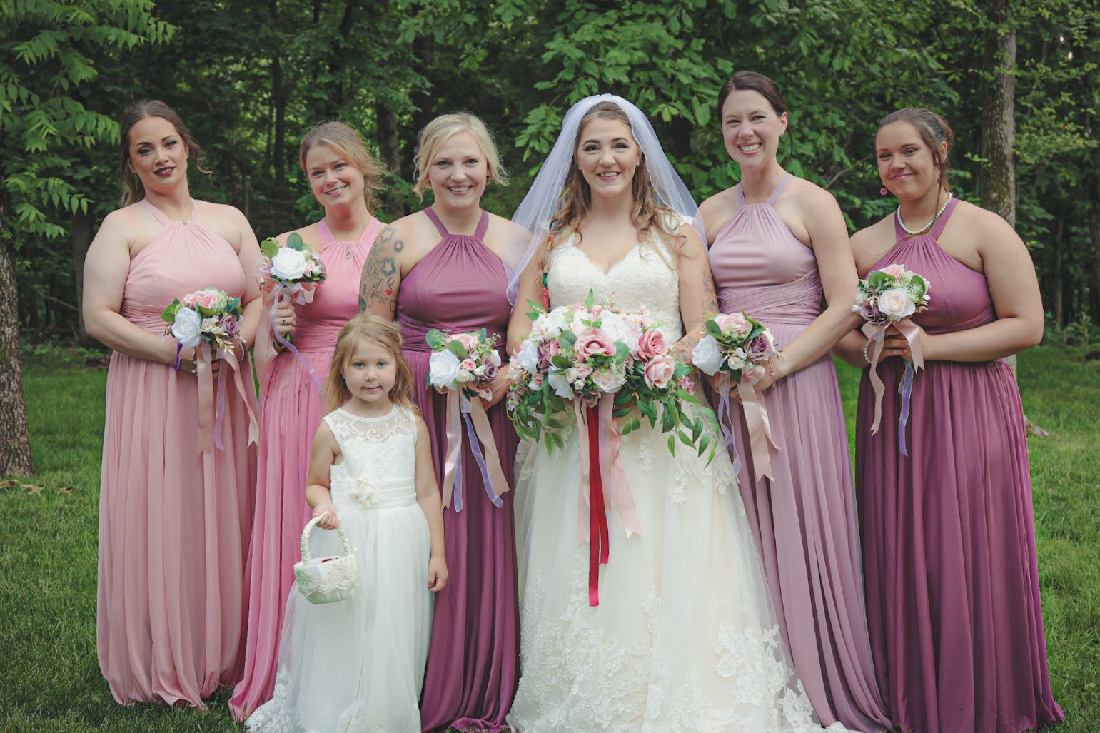 Wedding party in mauve and pink bridesmaid dresses poses together outdoors in a garden setting.