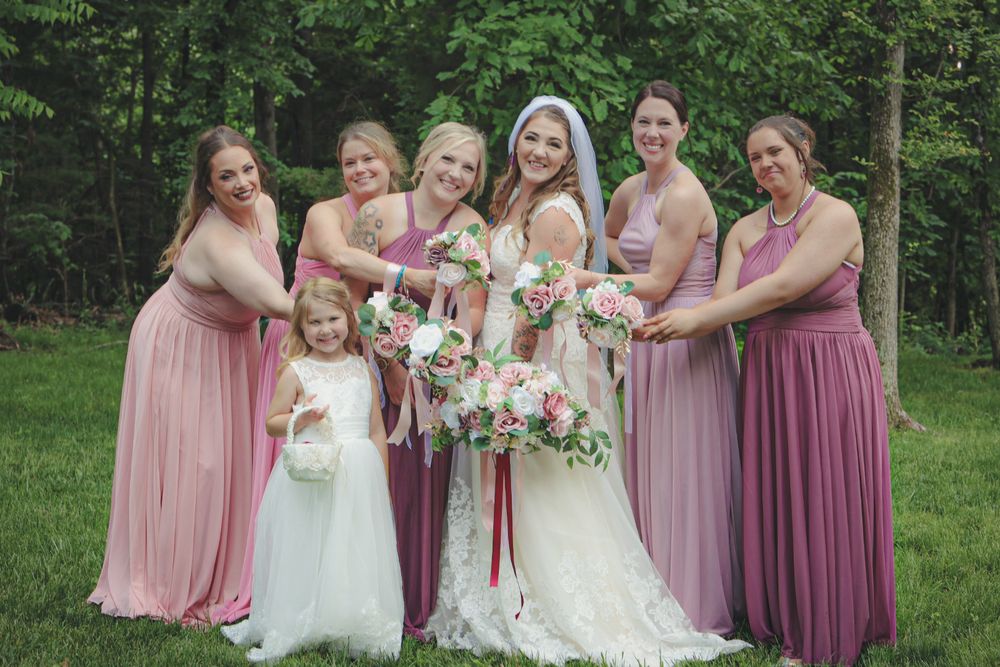 Bridesmaids in pink and mauve dresses pose together on green lawn during summer wedding.