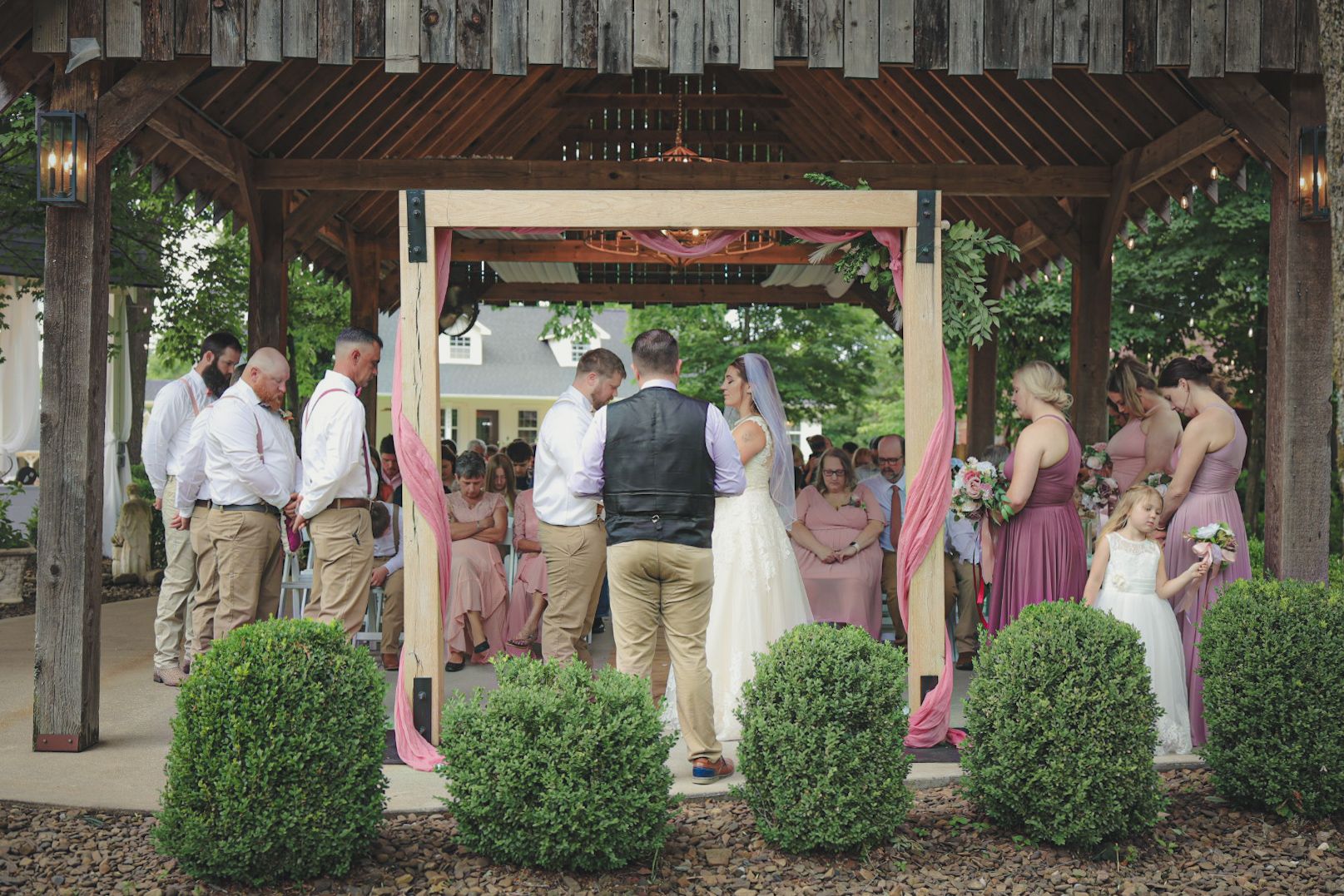 Wedding party gathers under a rustic wooden pavilion during an outdoor celebration.