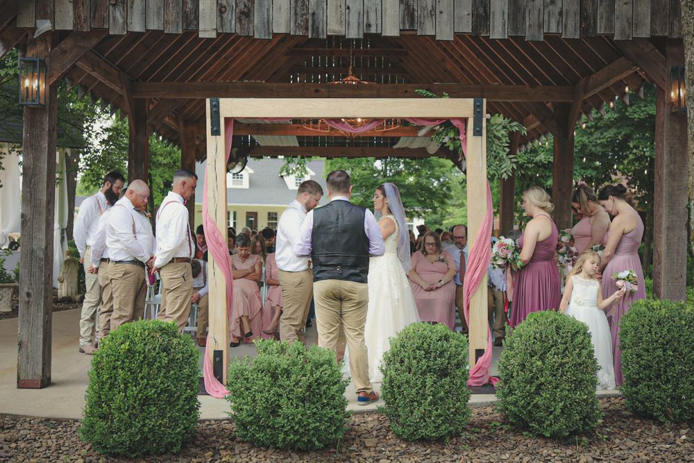 Wedding party gathers under a rustic wooden pavilion during an outdoor celebration.