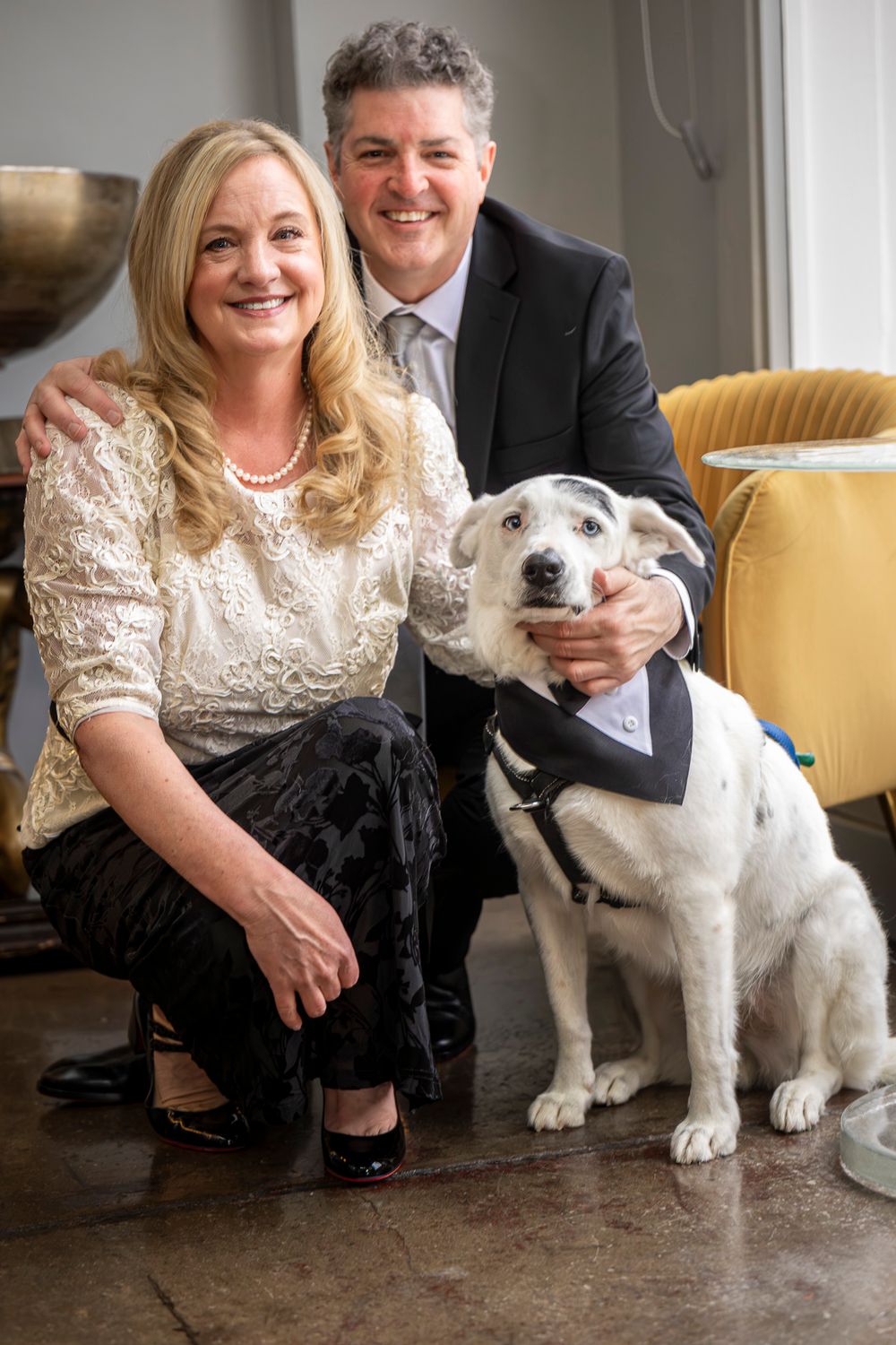 A white dog sits on a concrete floor next to people wearing formal attire.