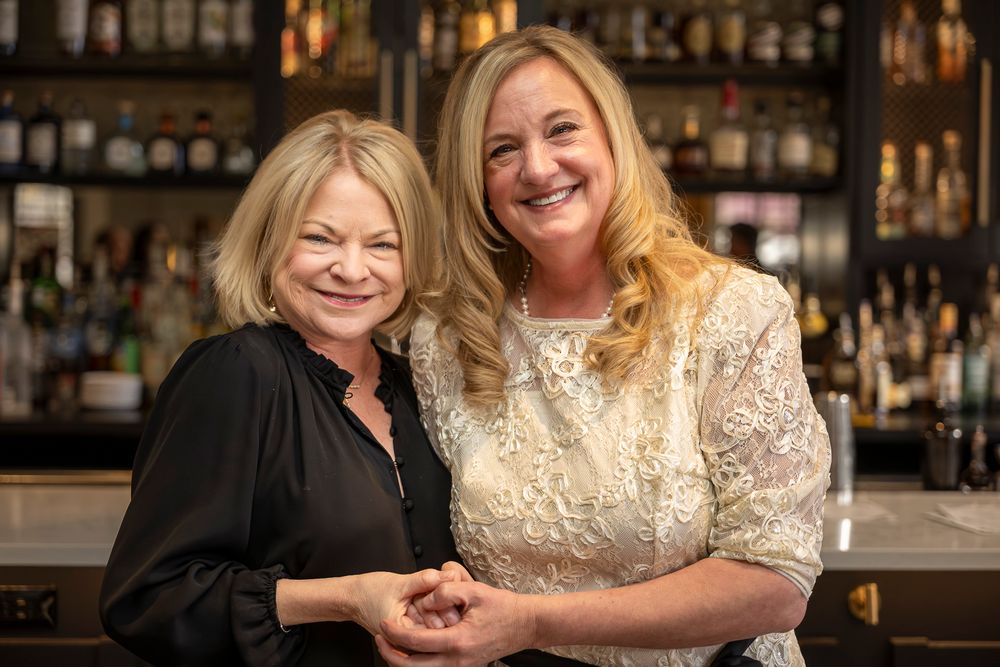 Two people smile warmly while sitting at a bar with bottles of liquor displayed on shelves in the background.
