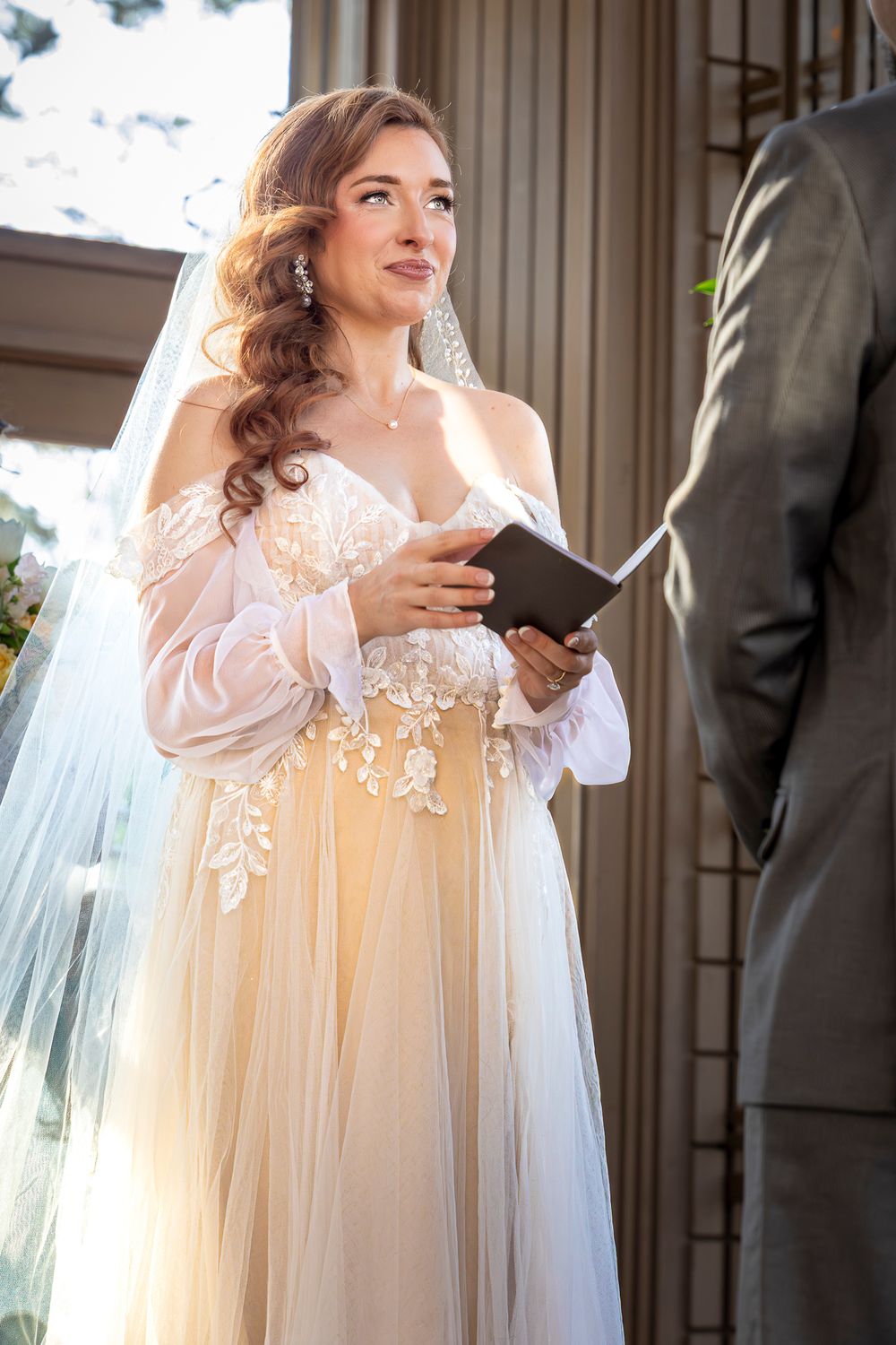 A bride in a flowing white lace gown and veil smiles while holding a mobile phone during a wedding ceremony.