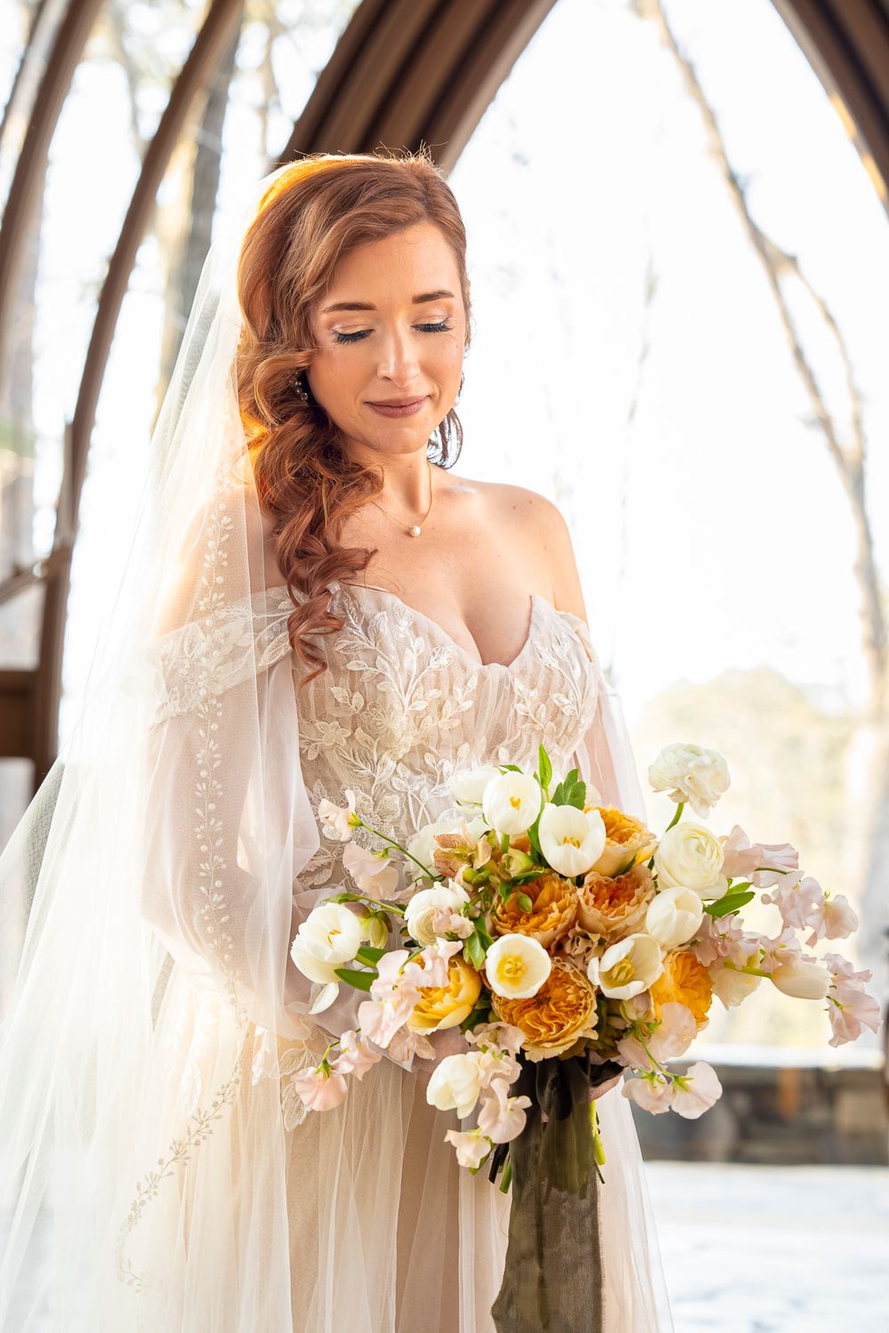 A bride in a lace gown holds a romantic bouquet of yellow and white flowers during a sunlit outdoor wedding ceremony.