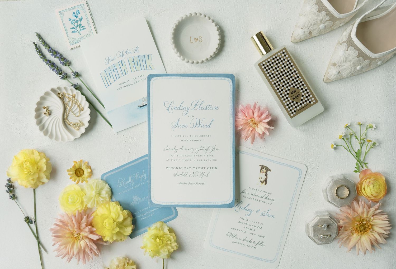 Wedding invitation suite with blue details surrounded by yellow and pink flowers and wedding accessories.