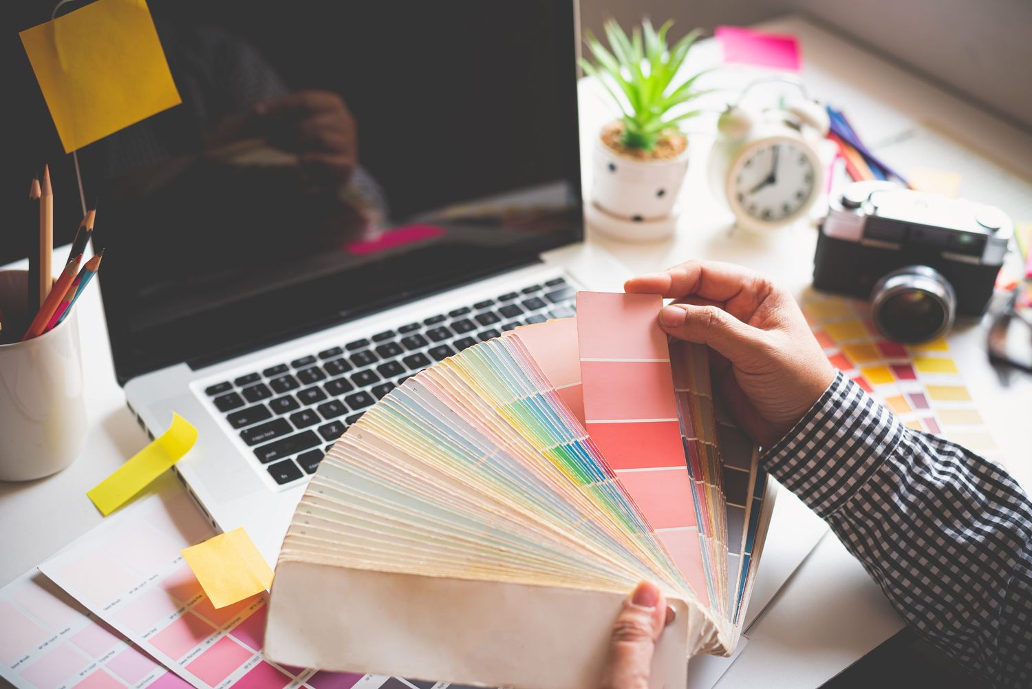 Designer reviewing color swatches at desk with laptop, coffee cup and small potted plant in workspace.