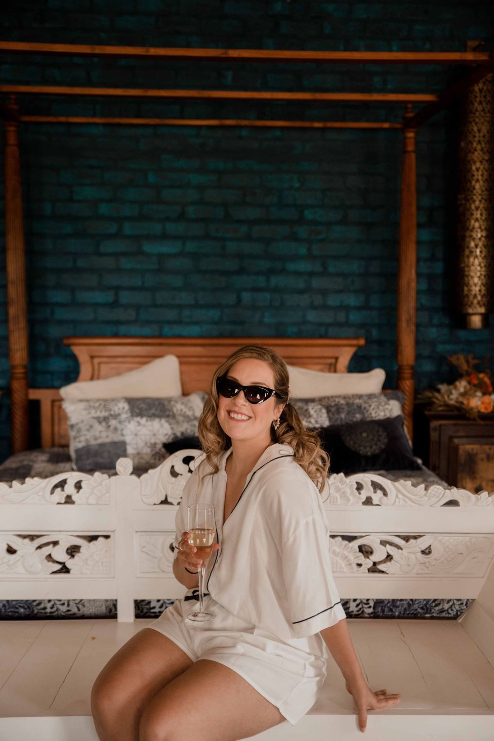 Bride posing with a glass of champagne in her wedding pajamas in front of a large four poster bed