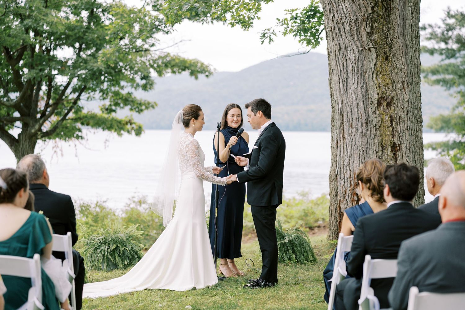 A wedding ceremony takes place outdoors under tall trees beside a scenic lake with mountains in the background.