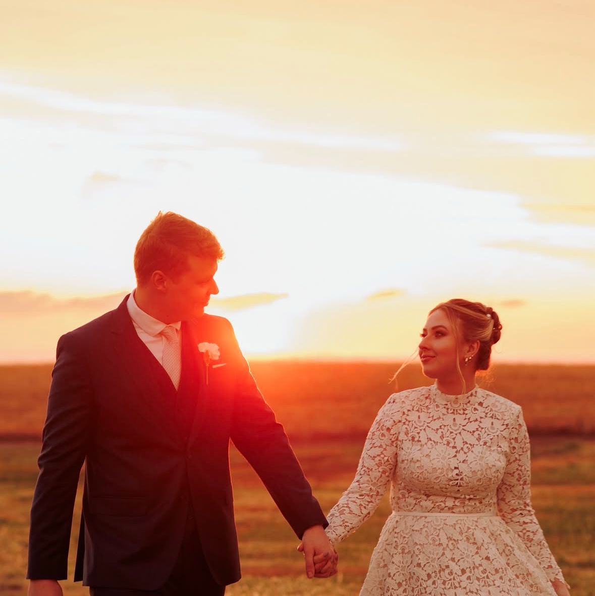 Silhouetted couple holding hands in lace dress and suit against dramatic sunset sky.