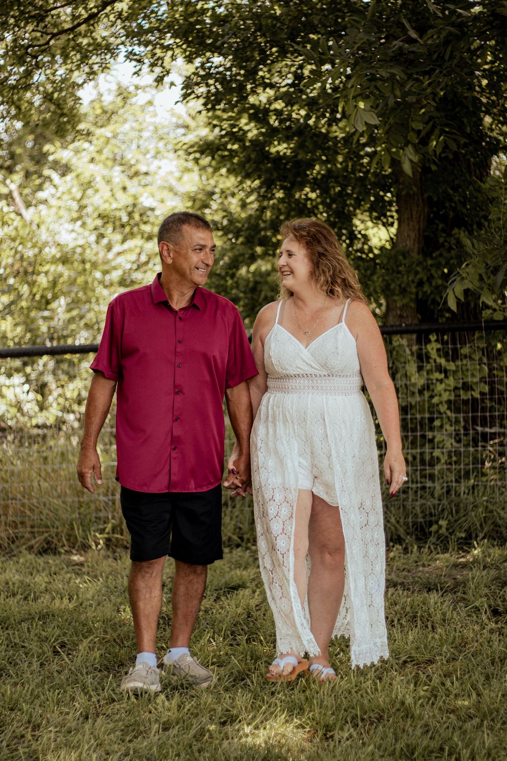 A couple walking in summer sunlight through a grass field wearing red and white outfits.