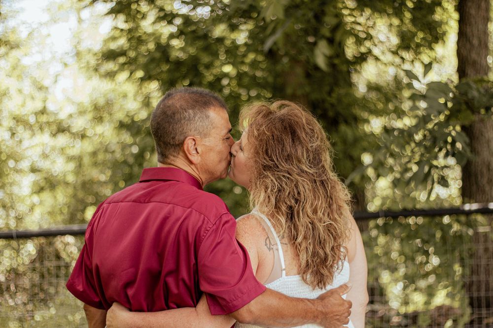 A couple in red and white embraces while sitting on a bench surrounded by lush green trees on a sunny day.