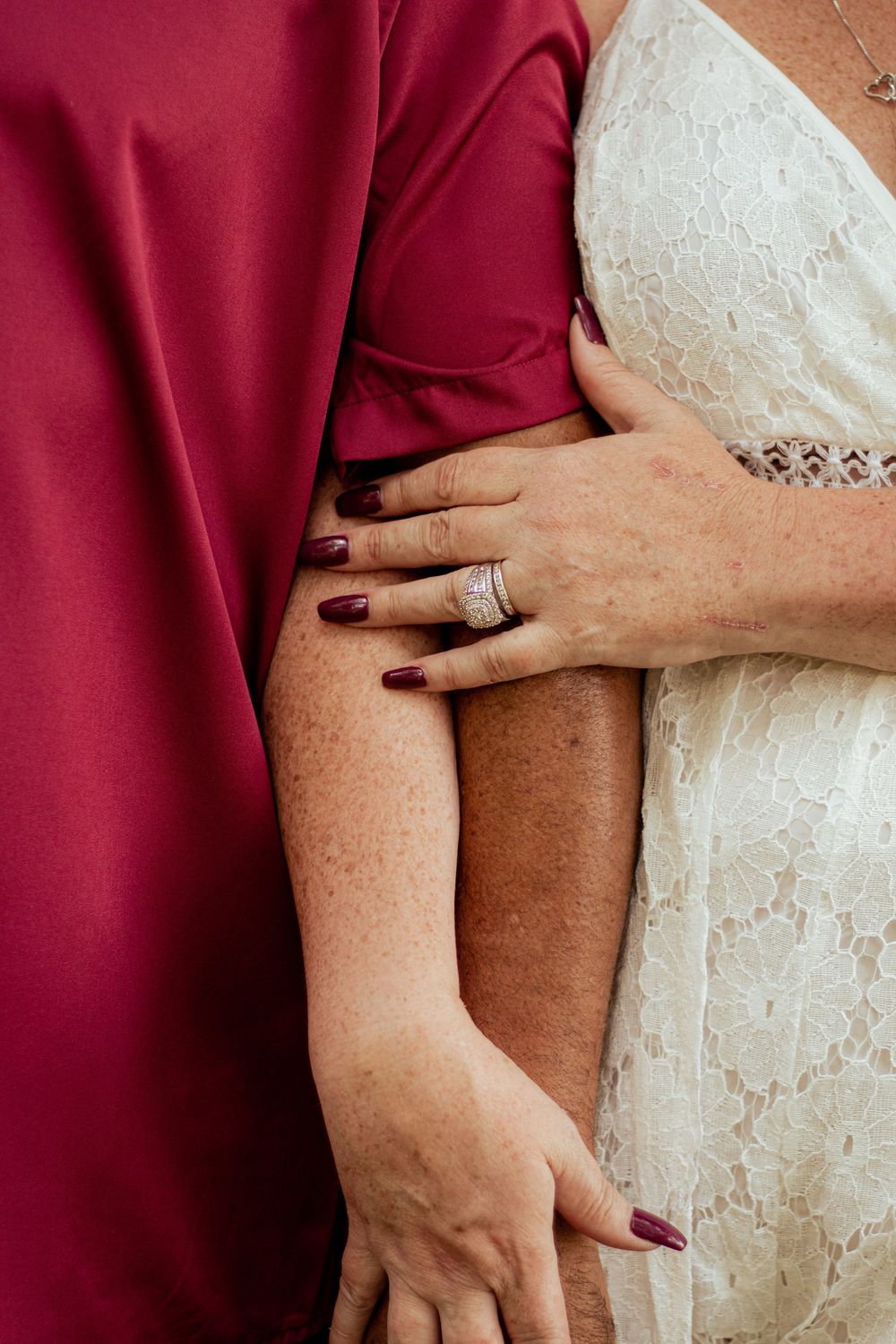 Two hands with burgundy nails wearing wedding rings touch against white lace and deep red fabric.