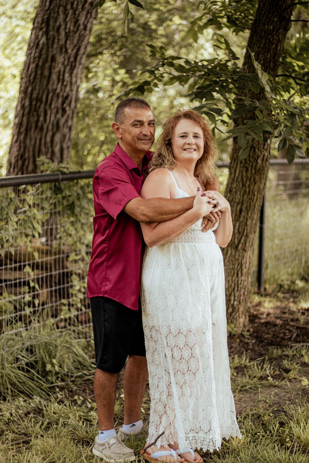 A couple embraces outdoors in a wooded setting, wearing a red shirt and white lace dress against a natural backdrop.