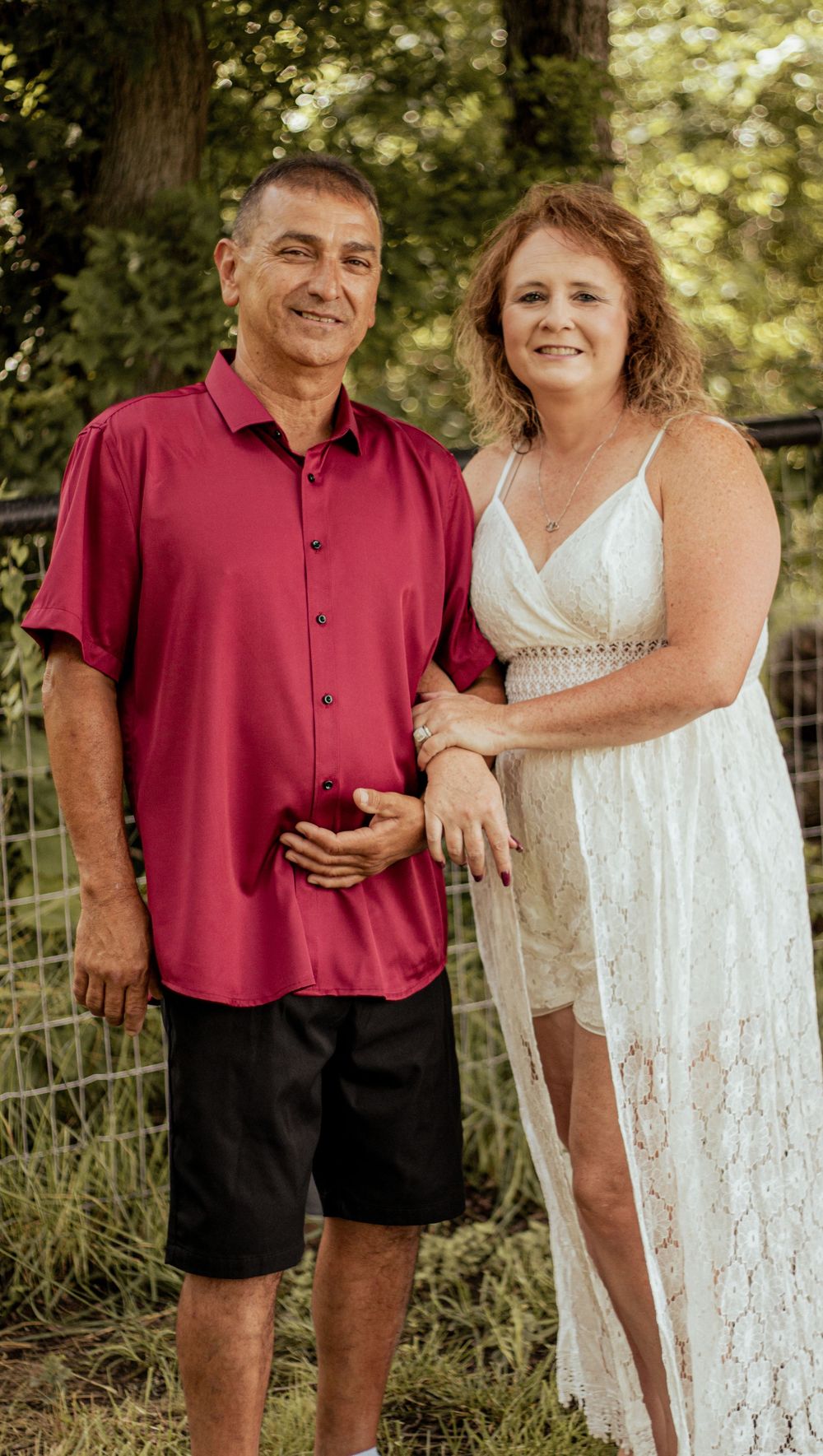 A couple poses together outdoors, with one wearing a red button-down shirt and shorts and the other in a white lace dress.