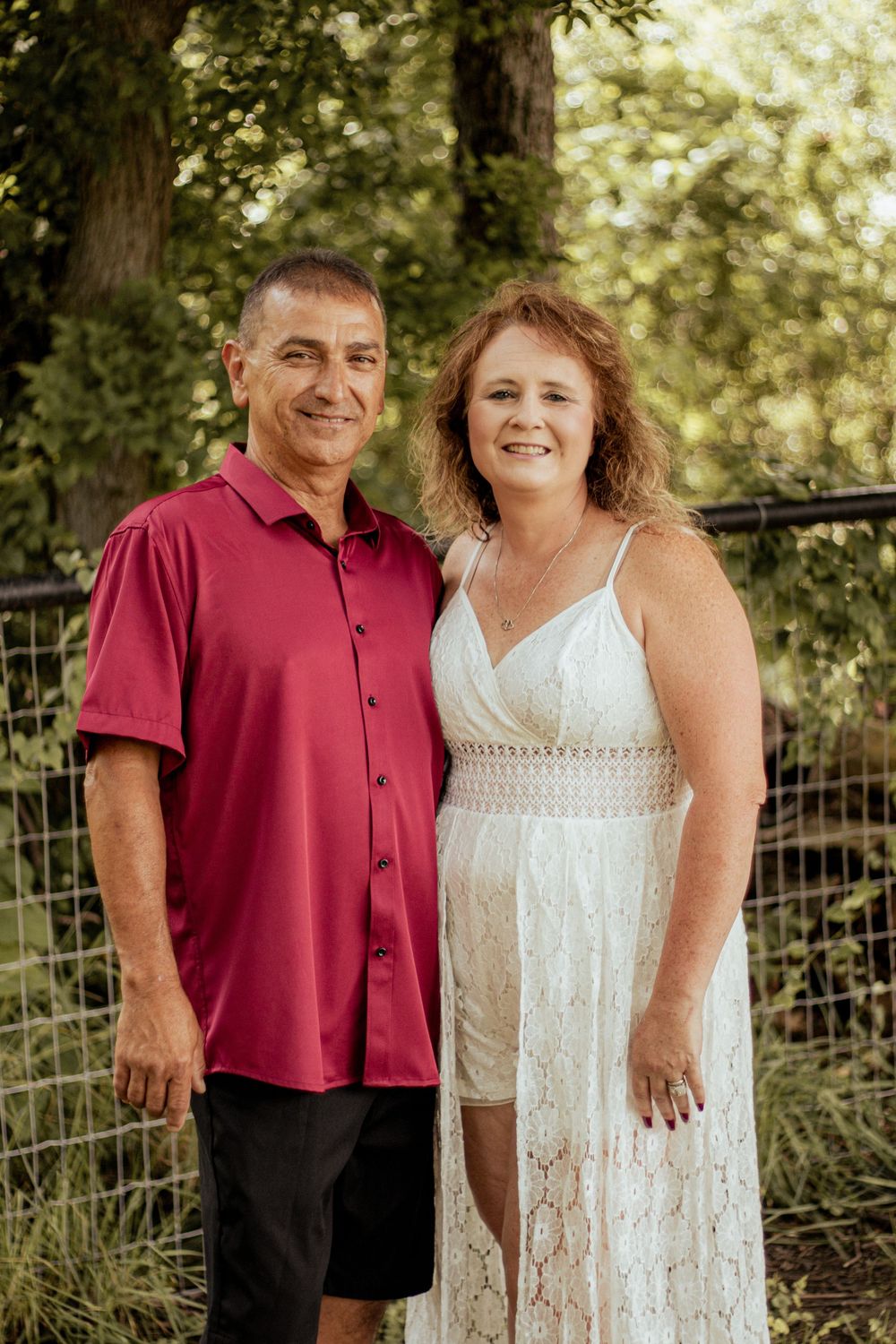 A couple poses together outdoors in summer attire, with a wooden fence and trees in the background.
