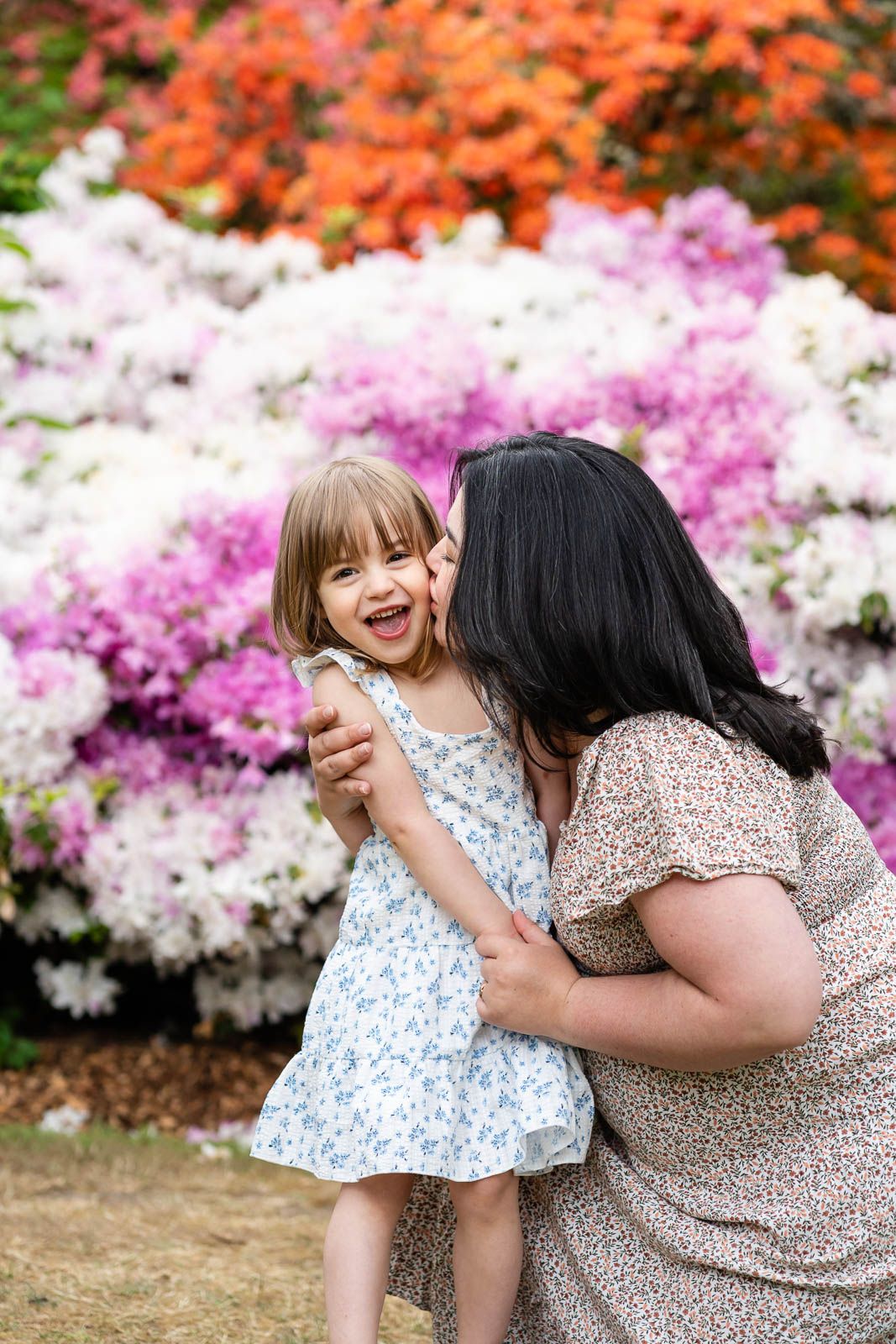Mom kissing daughter in front of rhododendron flowers.