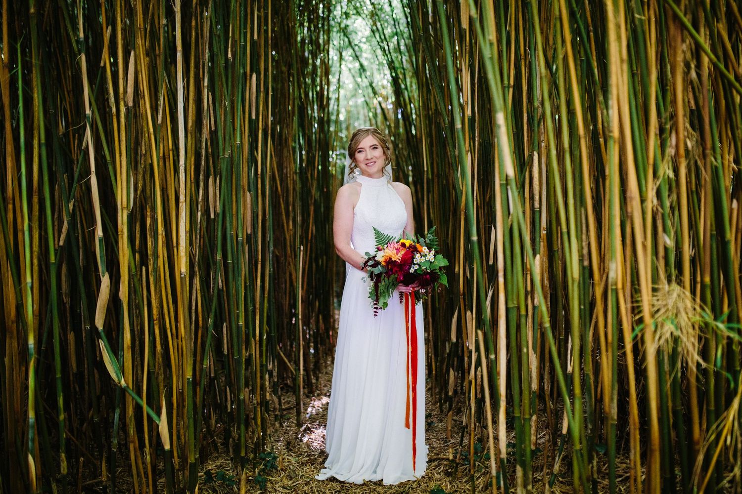 A bride in a white lace gown holds a colorful bouquet while standing in front of tall bamboo stalks in a garden.