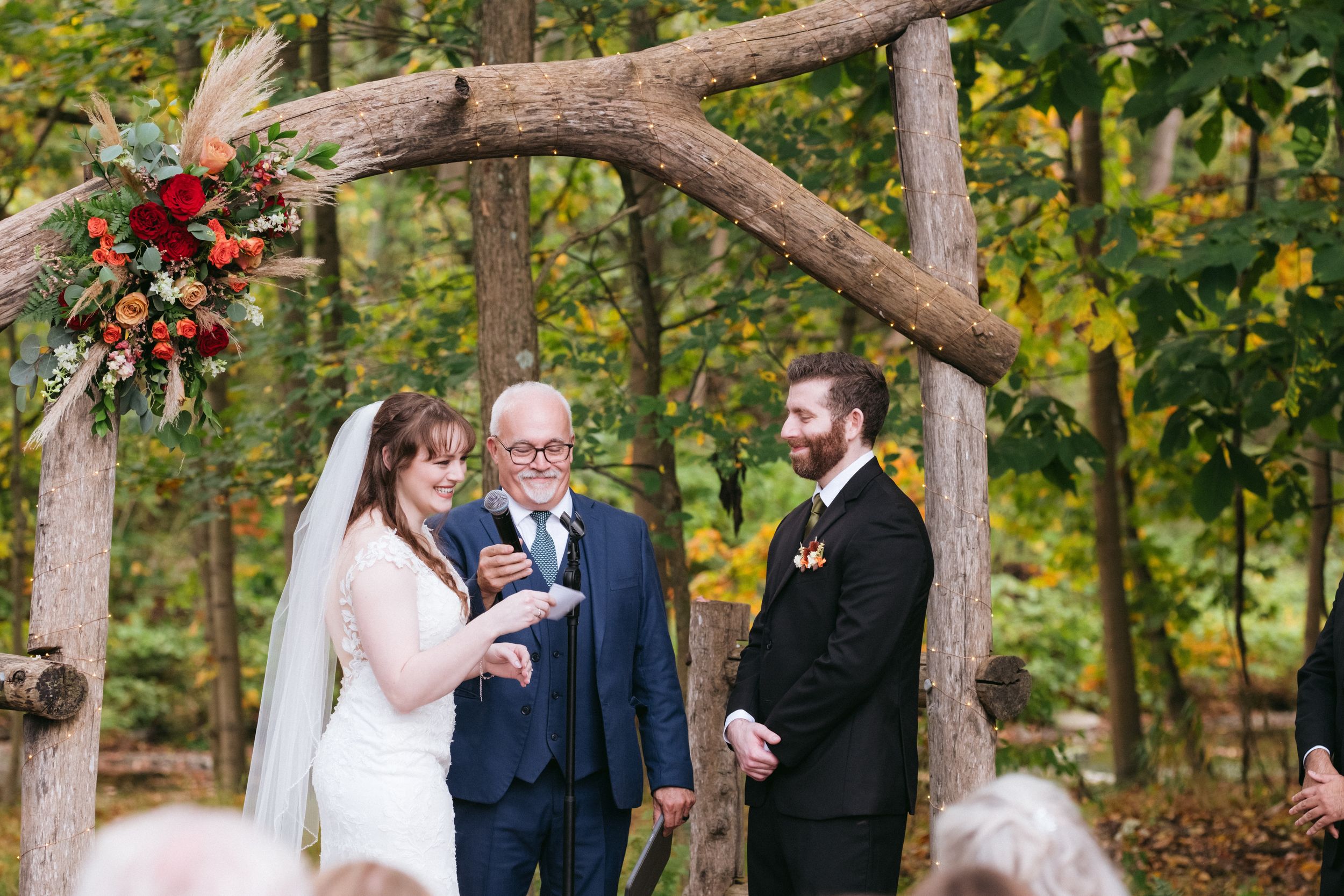 Outdoor wedding ceremony under rustic wooden arch with red floral decorations in autumn forest setting.