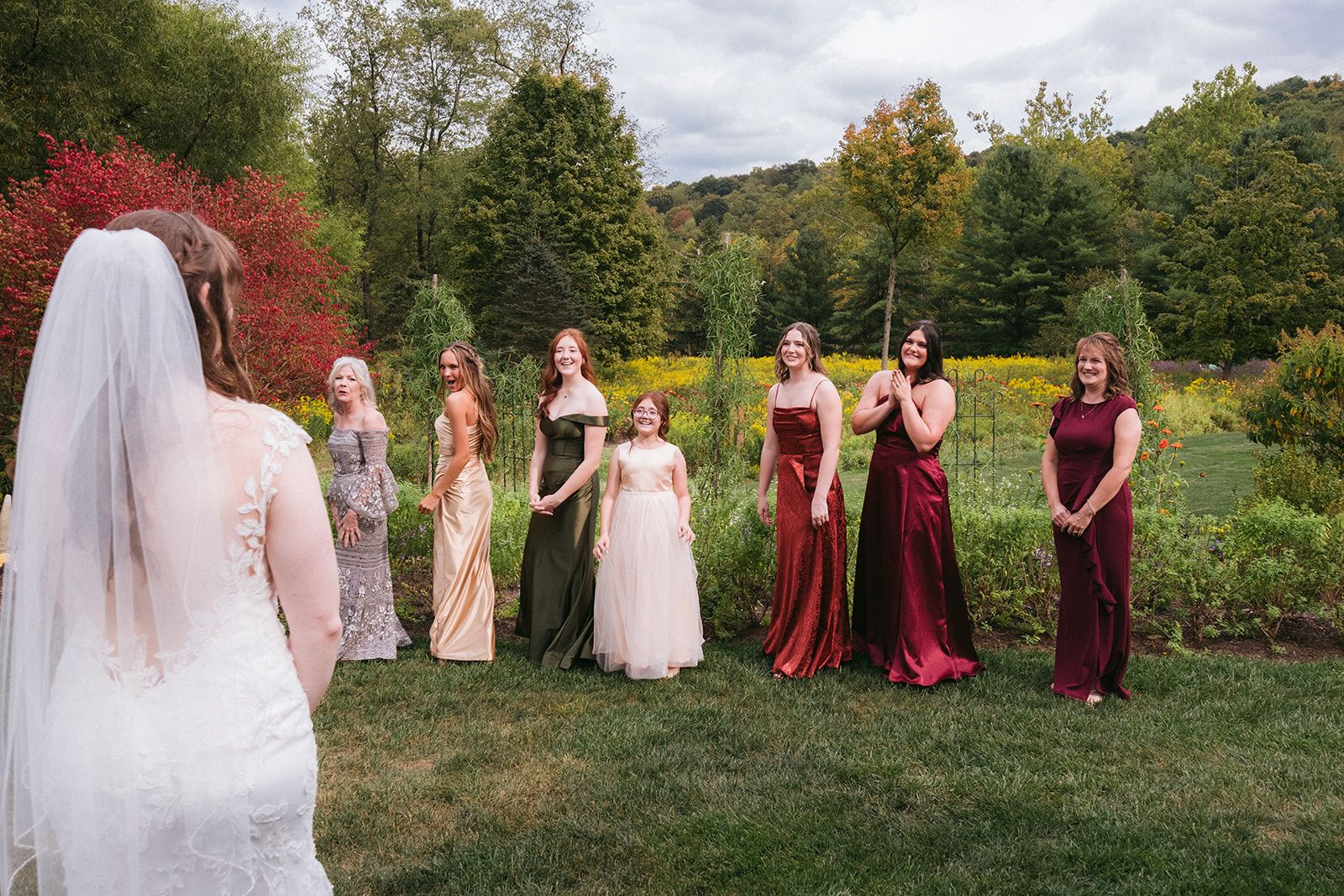 A bridal party stands on grass with autumn foliage in the background, featuring burgundy and blush colored gowns.