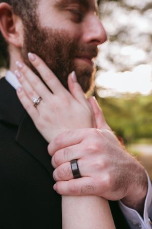 Close-up photo of hands with wedding rings as a bride touches the groom's bearded face in a tender moment.
