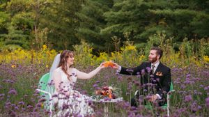 A couple toasts during an outdoor picnic surrounded by blooming purple wildflowers and forest greenery.