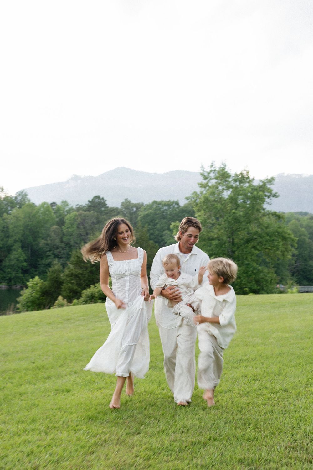 A family in white outfits runs joyfully across a grassy hill with mountains in the background.