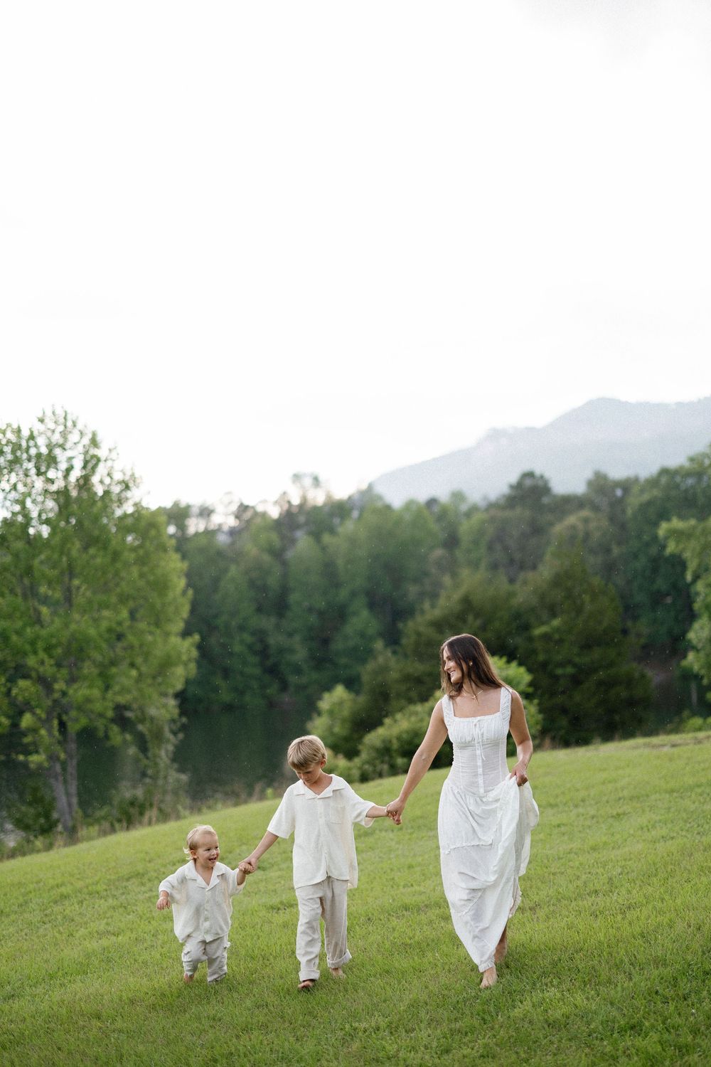 People in white dresses run playfully across a green hillside with mountains in the background.
