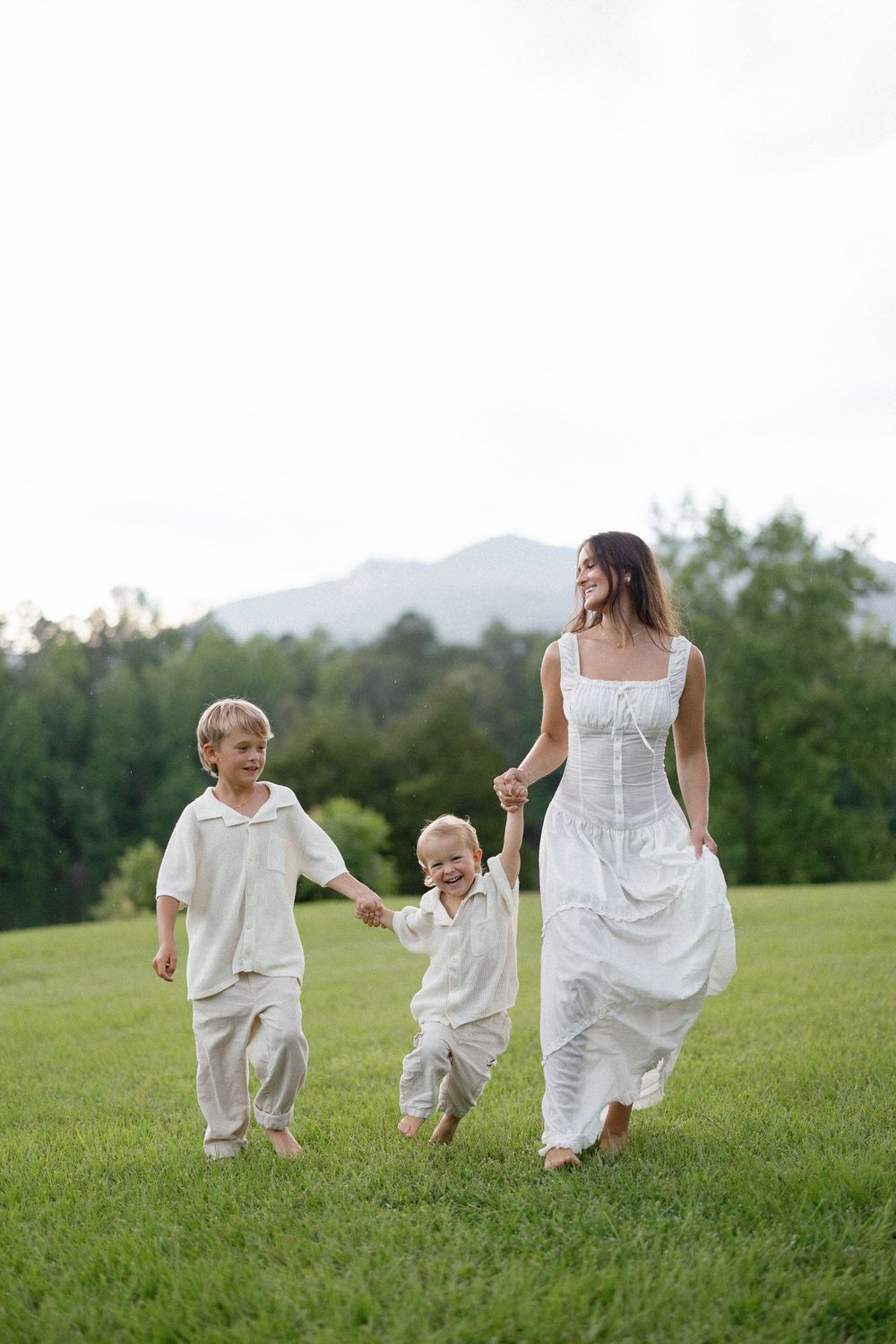 Two young children and a bride hold hands while running and laughing across a grassy hillside with mountains in the background.