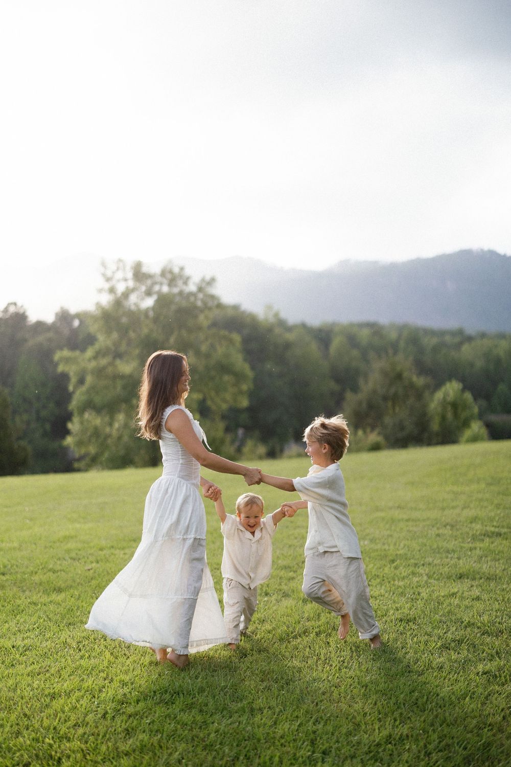 Three children in white clothing play and dance together in a lush green field with mountains in the background on a sunny day.