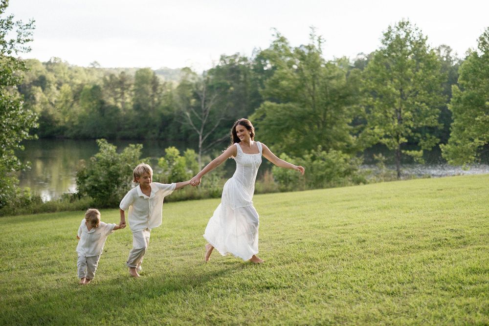 Happy couple in white wedding attire playfully chase children across a grassy hillside on a sunny day.