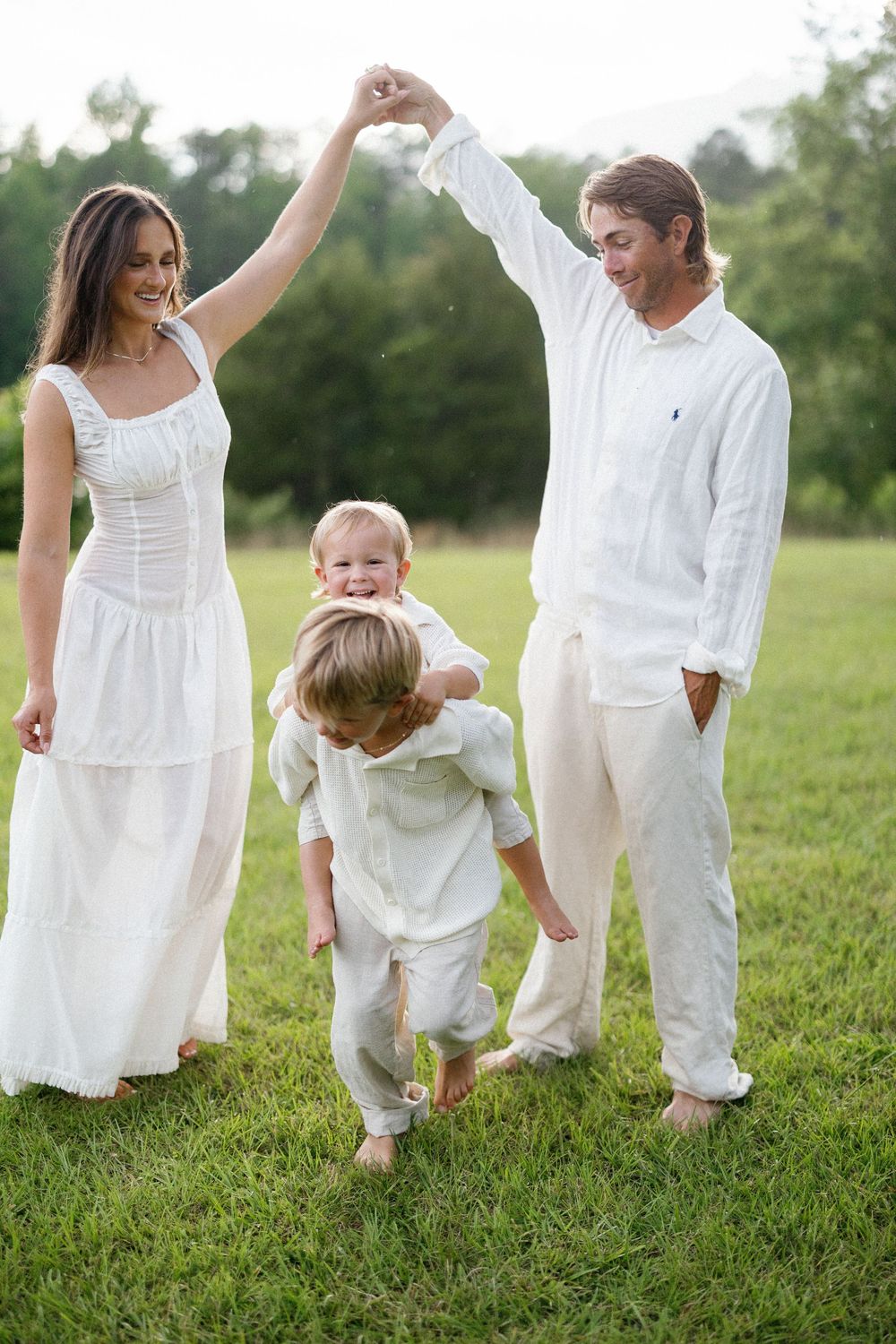 Family dressed in white outfits plays together in a grassy field on a summer evening, lifting and swinging their children.