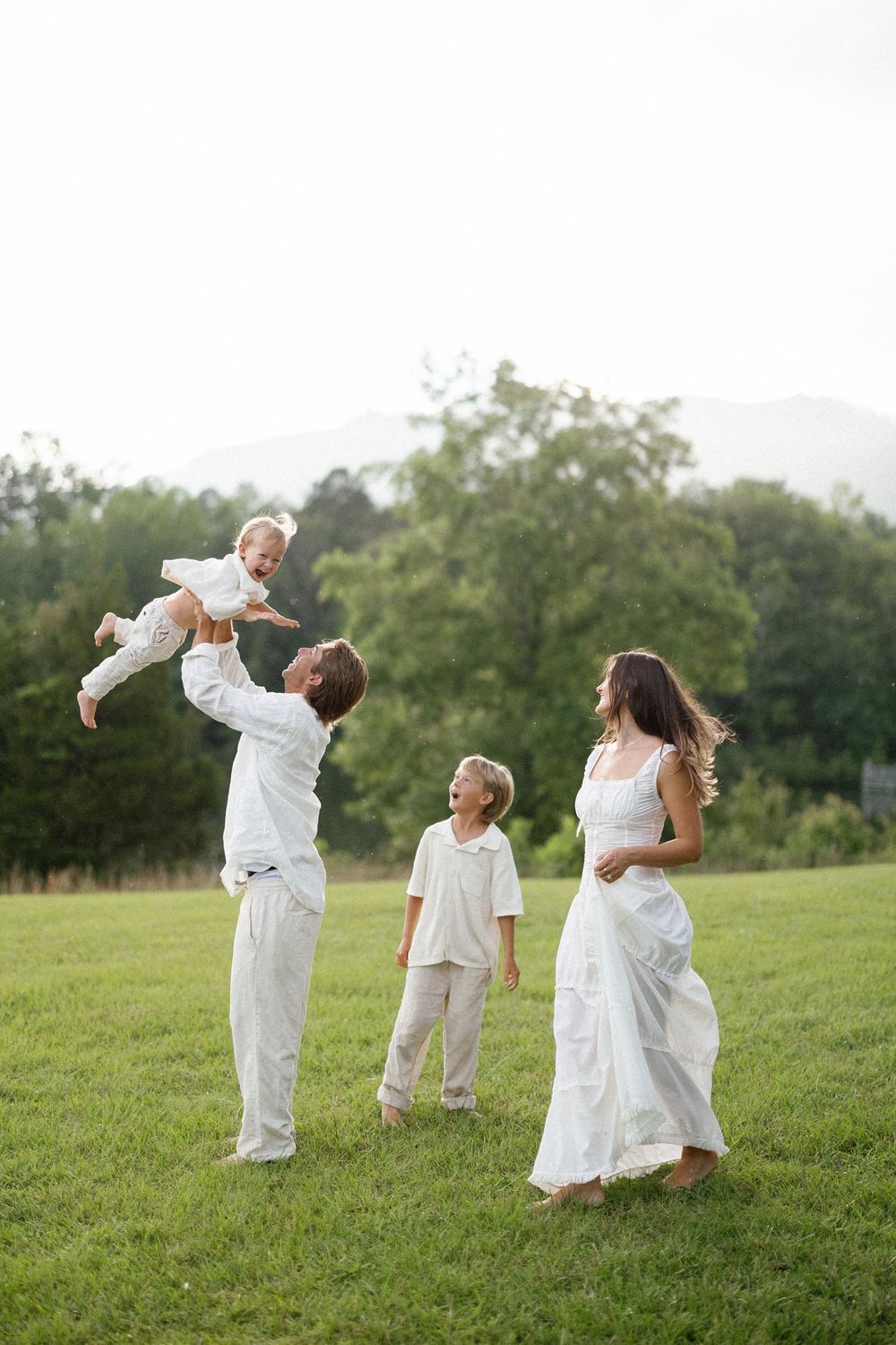A family in white outfits plays together on a grassy hill with trees in the background.