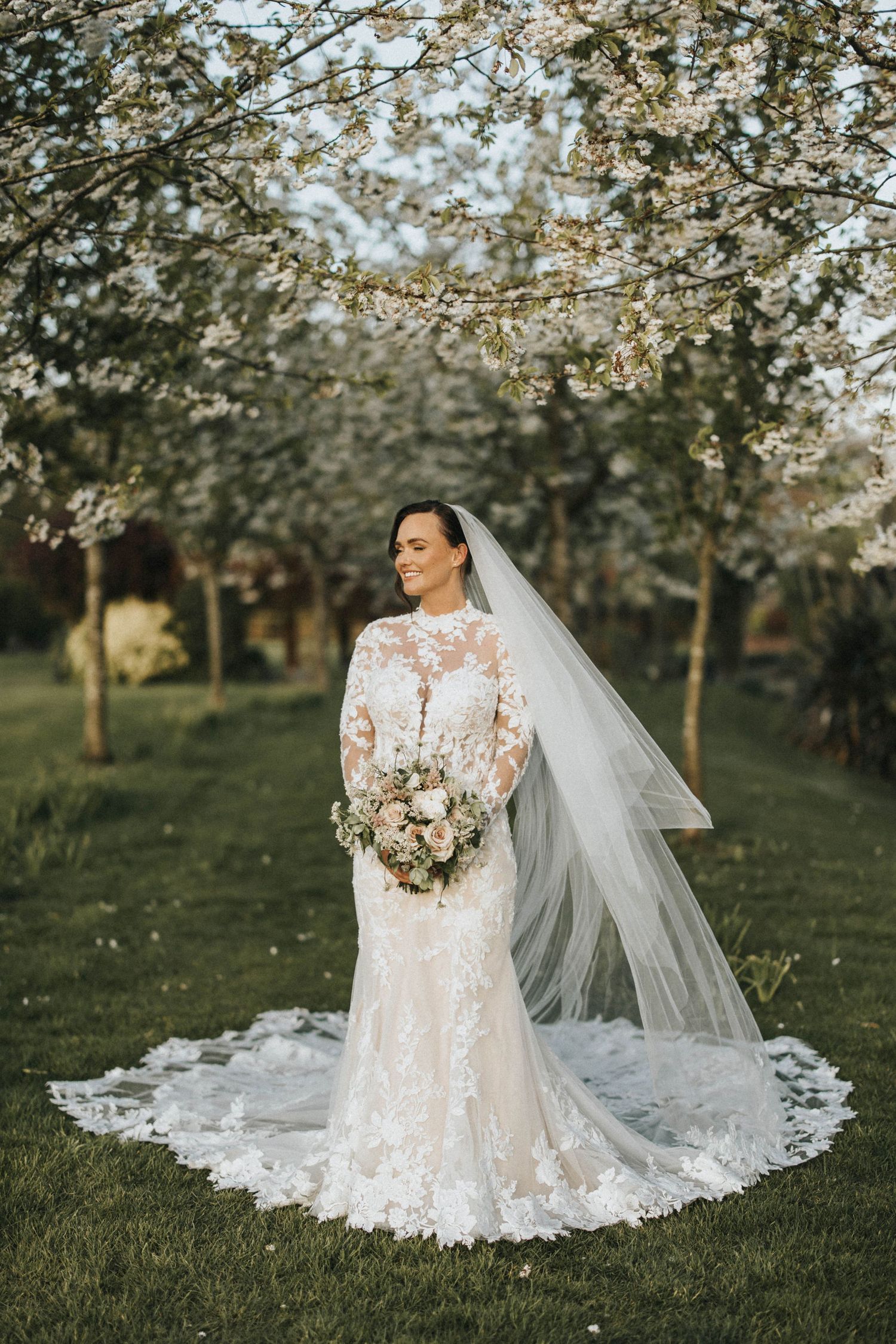 A bride in a flowing white lace gown with a long train stands beside a person in a peachy pink dress at night.