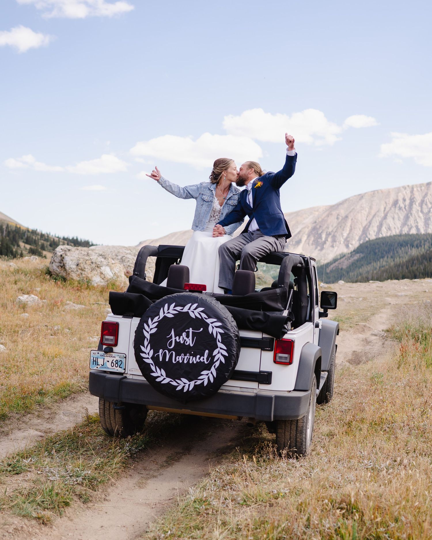 Newlyweds celebrating in a white Jeep Wrangler against mountain backdrop with 'Just Married' sign in rustic location.