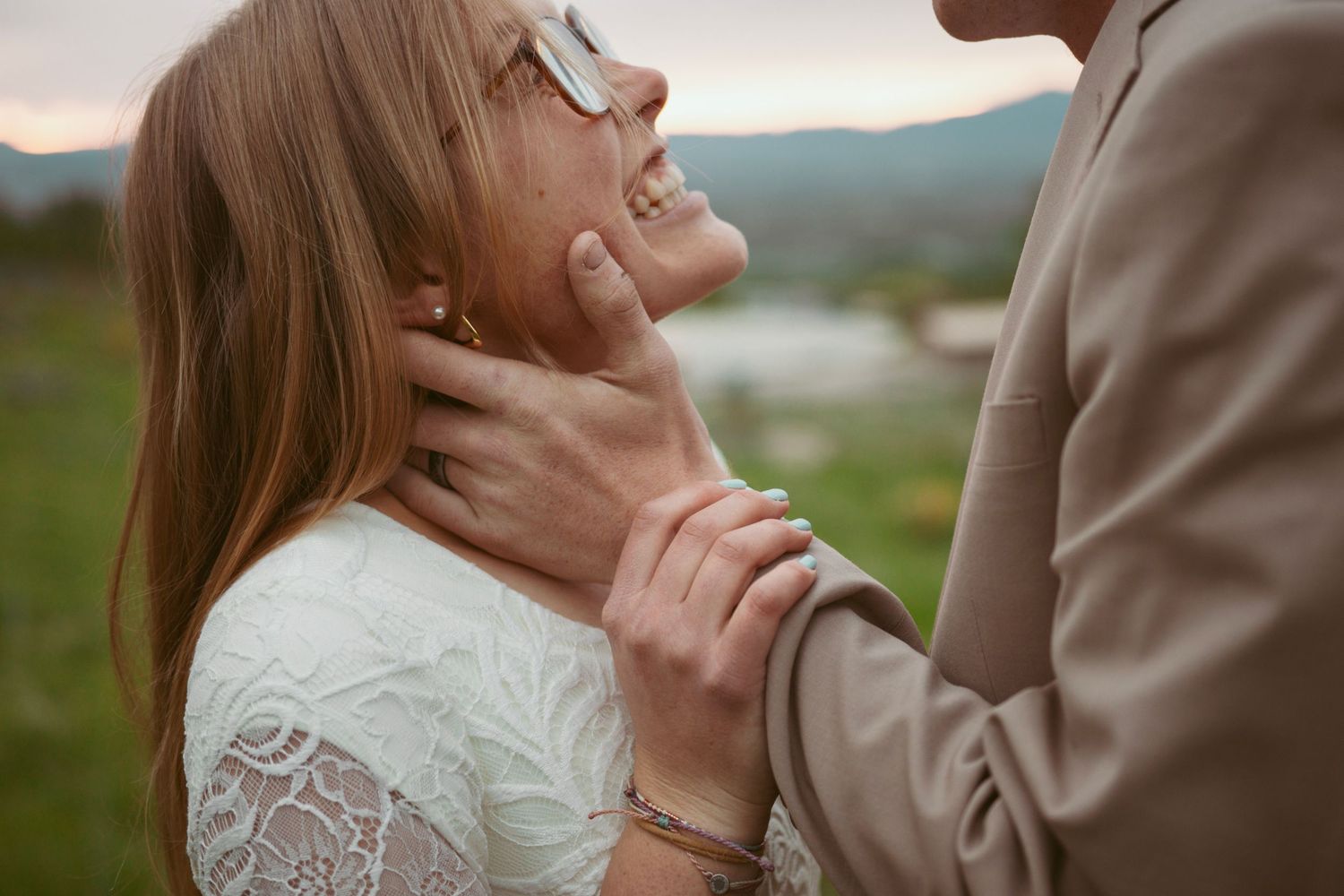 A couple stands in a sunlit meadow wearing wedding attire surrounded by evergreen trees during golden hour.