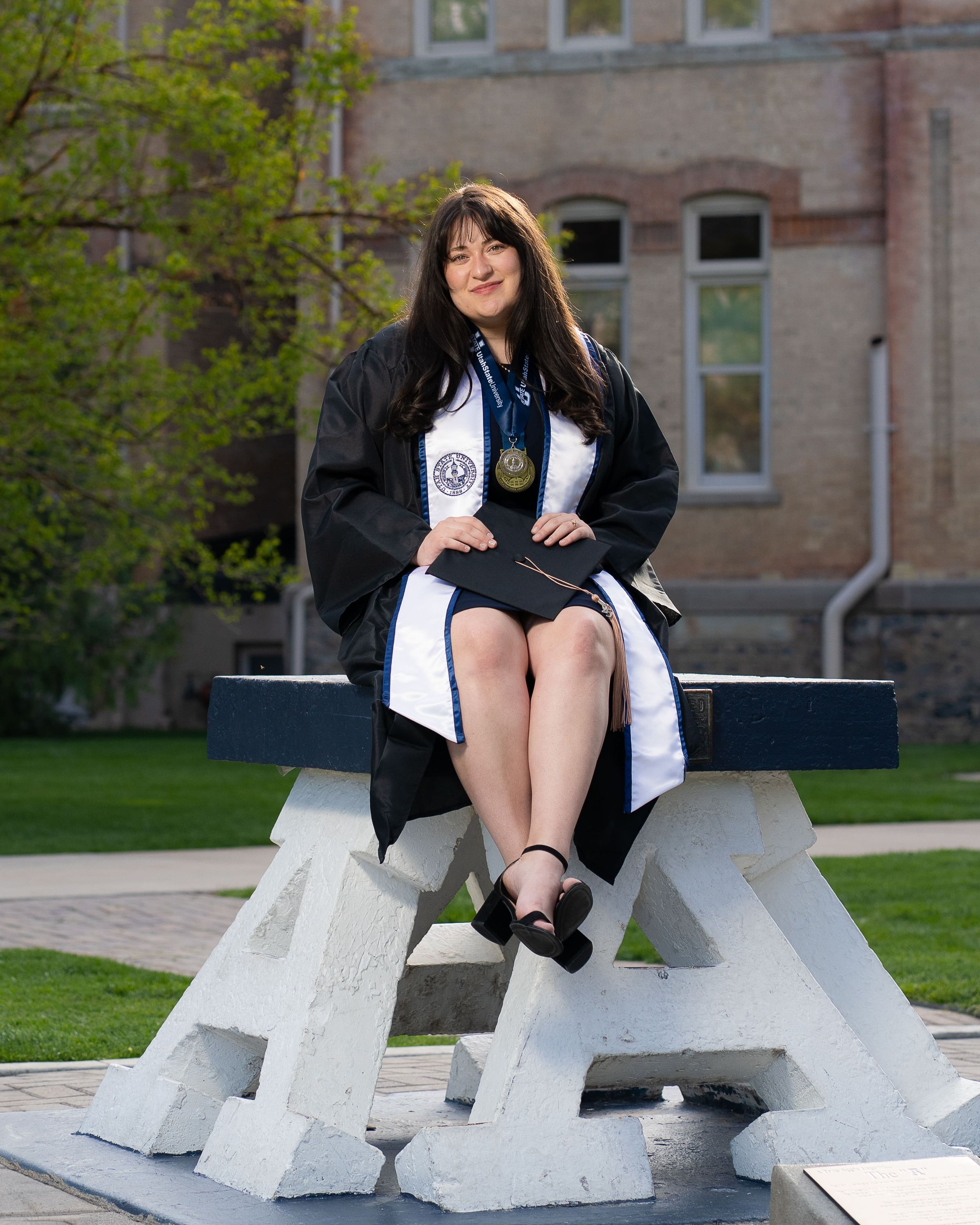 Utah State University female graduate in cap and gown sitting on USU "A" block in Logan UT.