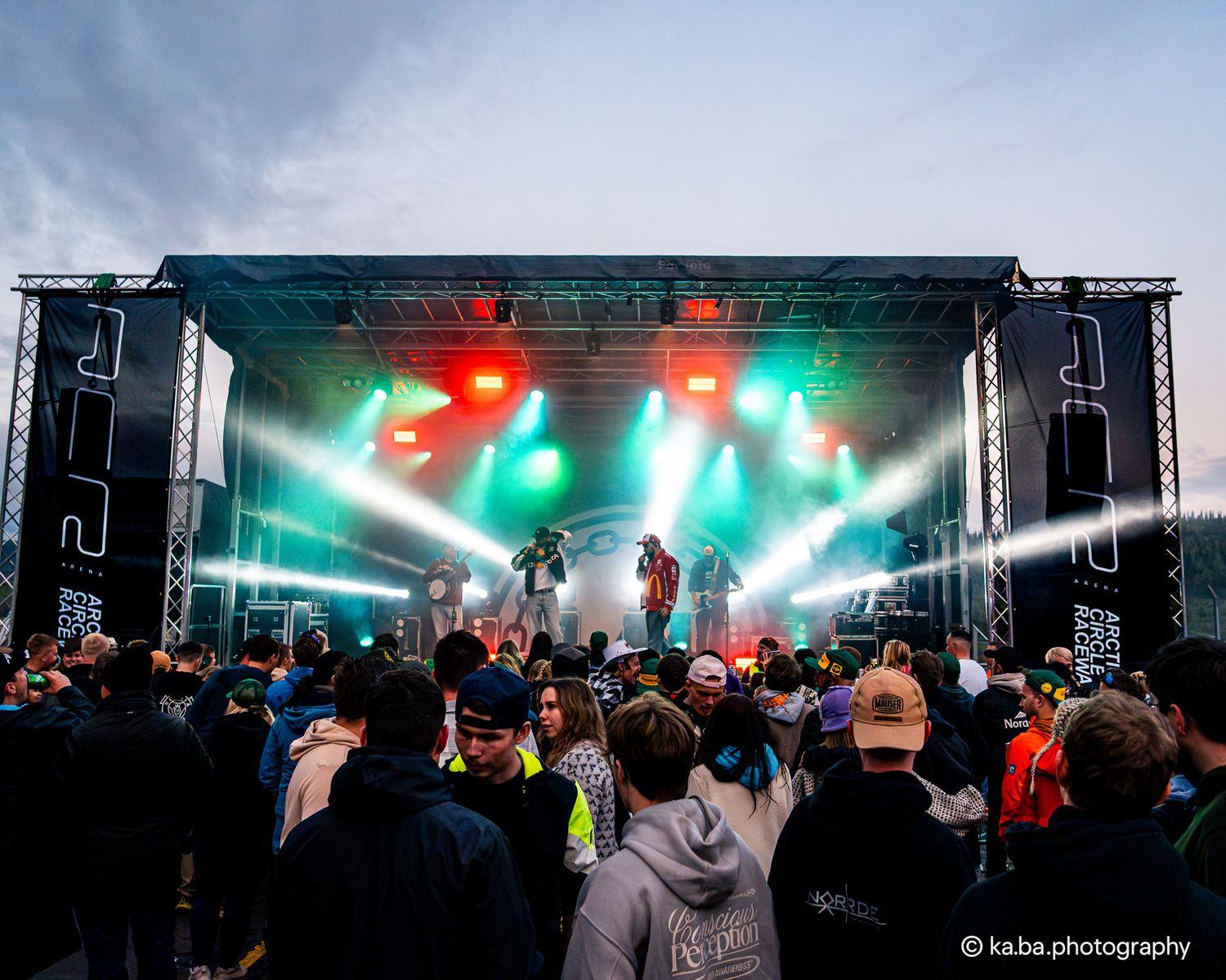 Outdoor music festival crowd gathered in front of a large illuminated stage with green and red lights at dusk.