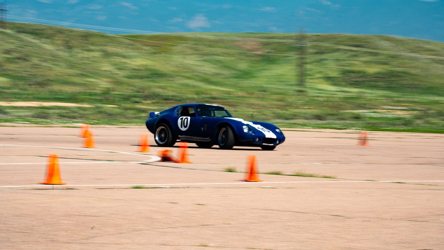 Black sports car speeds through orange traffic cones on a dusty autocross course with green hills in background.