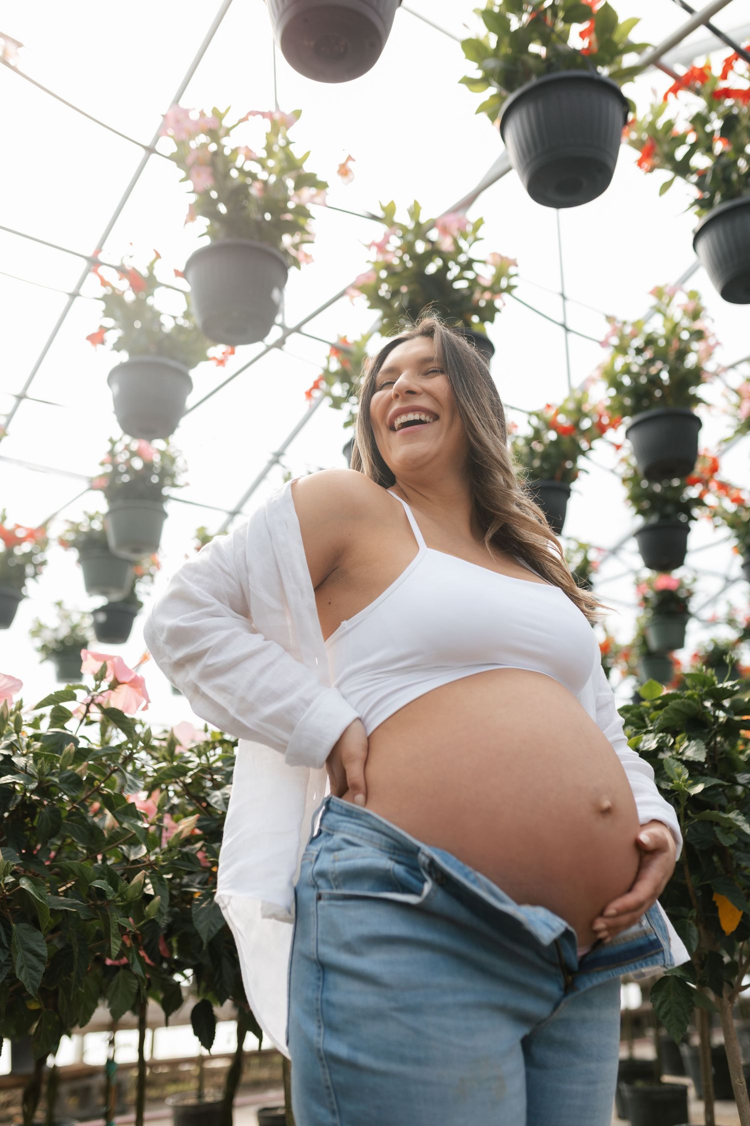 Smiling pregnant woman in white crop top and jeans cradling her baby bump in a lush greenhouse with hanging flower pots.